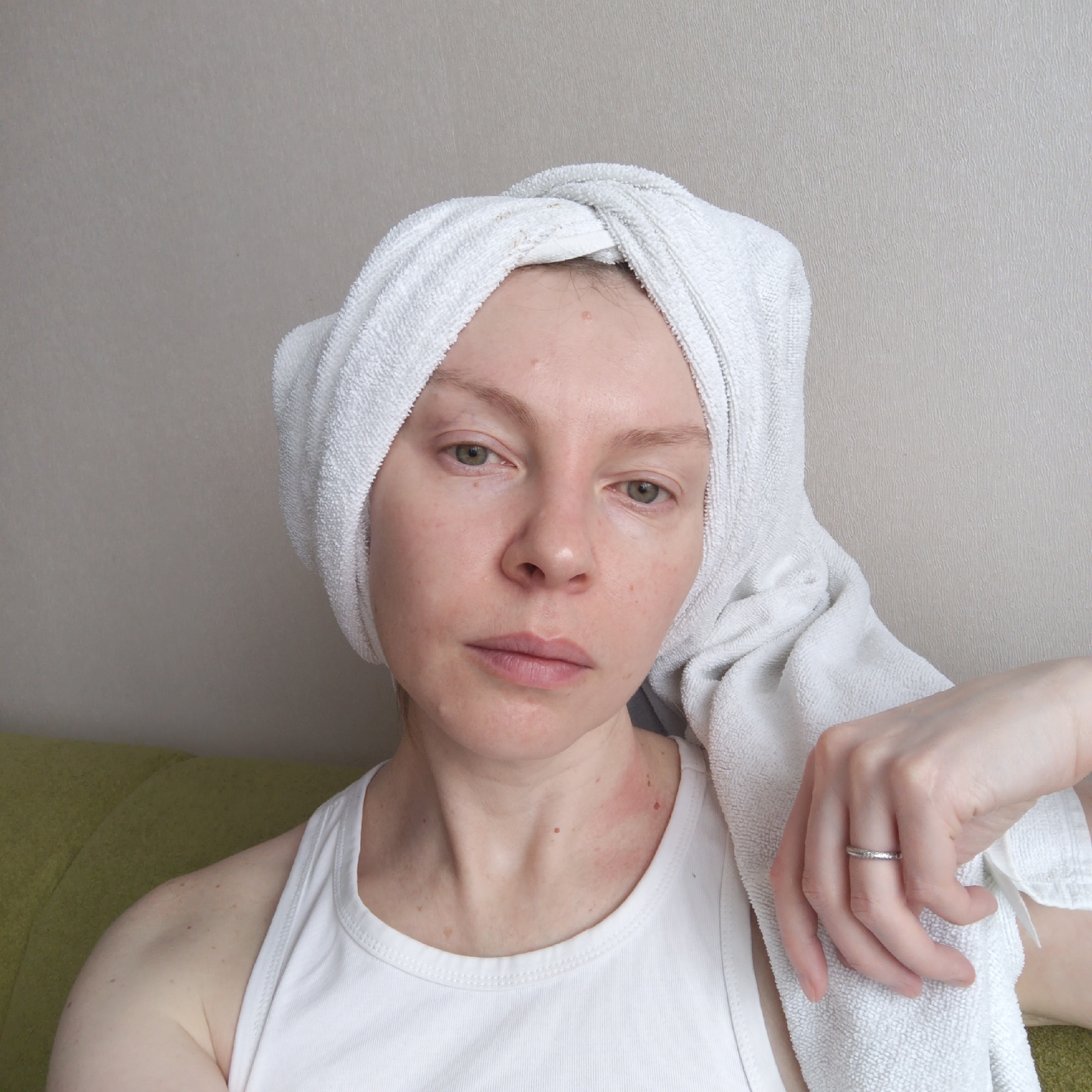 A pale-skinned woman with reddish undertones poses indoors with a white towel wrapped around her head. She wears a white tank top. Her hand is resting on the towel, and a simple ring is visible on her finger. Some redness is visible on her neck and face. The background consists of a plain wall and a green cushion behind her. The image was most likely taken in a domestic setting. There is no information available that connects the image to the city of Chaikovsky, Russia.