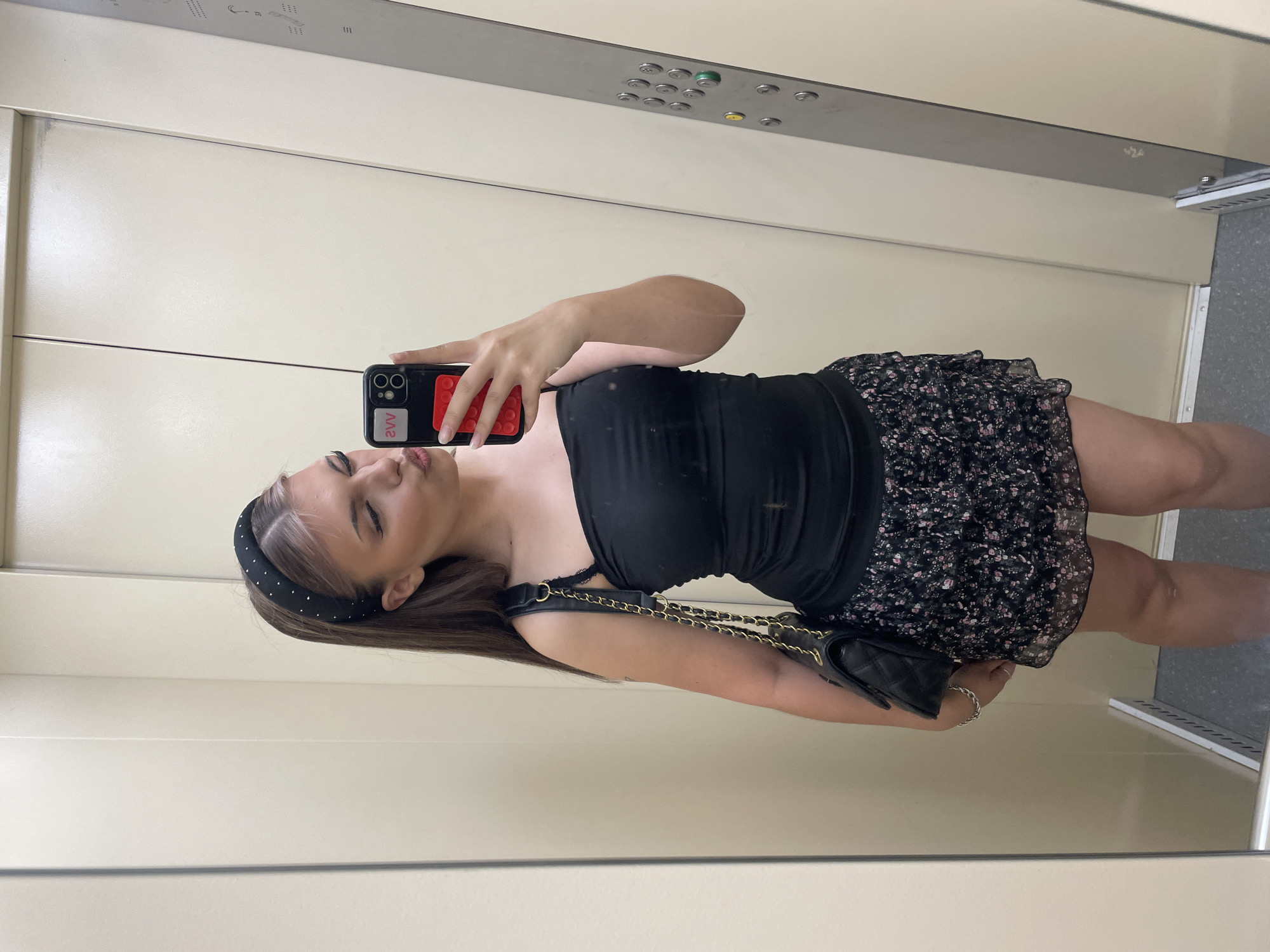 This media file captures a mirror selfie of a young woman inside what appears to be an elevator. The woman is the central subject, posing with her phone to take the photo. She is wearing a black strapless top and a floral print skirt. A black quilted handbag with a gold chain strap is slung over her shoulder. She has a padded headband adorned with small silver studs. Her expression is playful, with her eyes closed and lips pursed.
The background shows the interior of an elevator, with cream-colored walls and a metal panel displaying elevator buttons. The lighting suggests it could be daytime, but there are no strong indicators of natural light or specific weather conditions. The overall mood is casual and self-expressive, typical of a social media post.
There is visible text on the phone case, which reads "2VV" in white lettering on a black background. Part of a red object, possibly a Pop It toy, is also visible on the phone case. The elevator control panel has several circular buttons and indicator lights, one of which is green and another yellow. There is also some faint text on the metal panel above the buttons, appearing as numbers or symbols. The reflective surface of the elevator doors and walls creates a mirrored environment, doubling the visual elements.