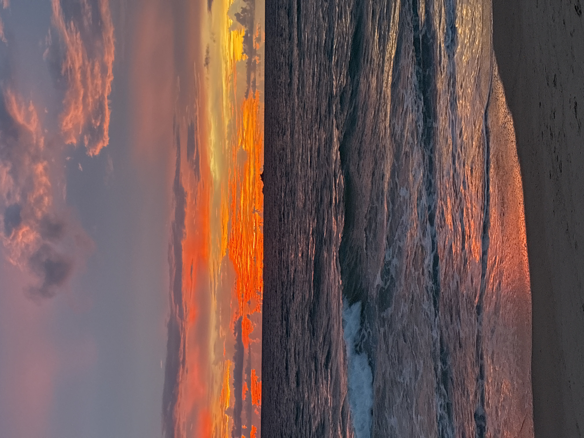 The image depicts a vibrant sunset over the Andaman Sea, viewed from a beach in Choeng Thale, Thailand. The sky is ablaze with fiery oranges, reds, and yellows, with streaks of pink and purple hues from scattered clouds. The sun is low on the horizon, casting a warm glow across the scene. The ocean is a deep, dark blue, with gentle waves rolling towards the shore. The water reflects the colors of the sunset, creating a shimmering effect. A small boat or distant landmass is visible on the horizon. The foreground shows a sandy beach, with the wet sand near the water's edge reflecting the sunset colors. There are no people or animals visible in the image. The scene conveys a sense of tranquility and natural beauty.