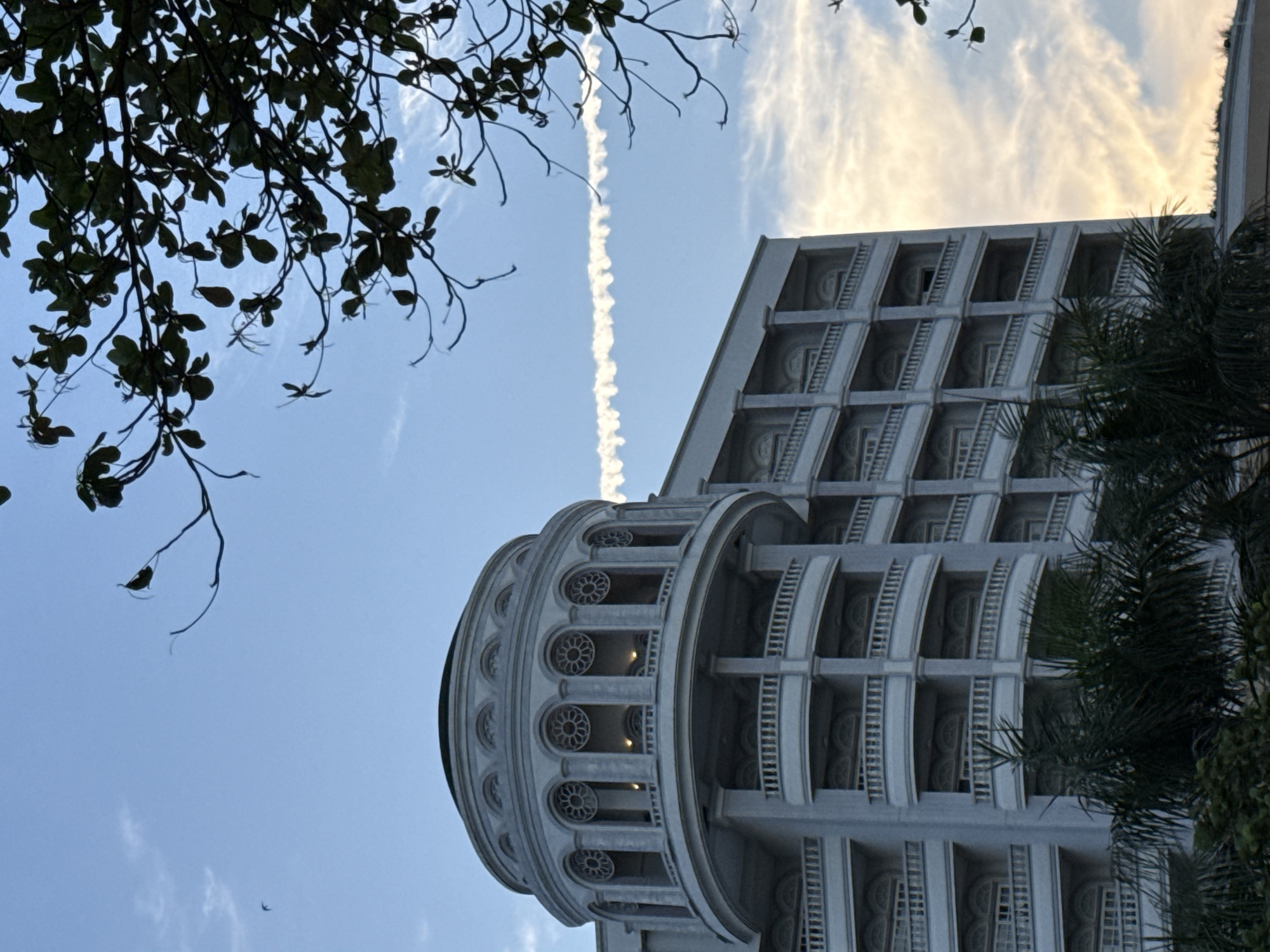 The image depicts a detailed view of a multi-story building with a domed structure on top, likely located in Nha Trang Ward, Vietnam. The architecture is ornate, featuring arched windows with decorative elements, multiple balconies with railings, and classical columns. The building's facade is light-colored, possibly white or light gray. In the foreground, lush green palm fronds and other foliage partially obscure the lower levels of the building. The sky above is a light blue, with wispy white clouds and a distinct contrail stretching horizontally across the frame. A few branches of a tree with dark green leaves are visible in the upper right corner, silhouetted against the sky. The lighting suggests it might be late afternoon or early morning, given the soft illumination and the colors of the sky. There are no people visible in the image.