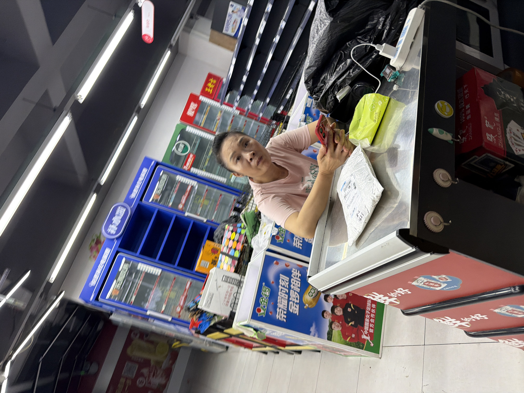 A woman is sitting at a metal counter in a convenience store in Jiangjin, China. She is holding a mobile phone in her right hand and has a package on the counter in front of her. There are various items on the counter, including a power strip, a charging cable, and a green packet of tissues. In the background, there are shelves stocked with products and a display of soft drinks. The store appears to be well-lit, with fluorescent lights on the ceiling. The overall impression is of a typical retail environment.