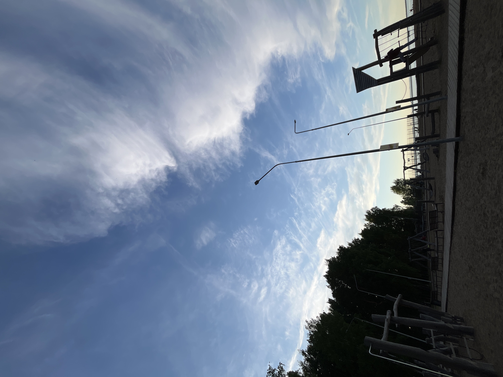 The image captures an outdoor recreational area, likely a sandy beach or park situated beside a large body of water, under a bright, partly cloudy sky. The perspective is slightly upward-angled, emphasizing the expansive sky.
The upper portion of the frame is dominated by a vibrant blue sky, adorned with sweeping formations of white, wispy clouds, resembling cirrus. These clouds are more dense and concentrated on the left side, gradually thinning out to reveal clearer blue sky towards the right.
In the foreground and mid-ground, a sandy expanse stretches across the lower part of the image. Scattered across this sandy terrain are various outdoor exercise or playground structures, constructed from wood and metal. These include climbing frames, balance beams, and other fitness equipment, appearing largely as dark silhouettes against the brighter sky and distant water. Two tall streetlights, characterized by their curved arms, also rise from the sandy ground, prominently silhouetted against the sky. A dense line of dark green trees forms a natural boundary along the right side of the recreational area, their foliage similarly silhouetted.
In the distant background, a calm body of water is visible, reflecting the ambient light. Beyond the water, a faint and indistinct shoreline or distant structures can be perceived. The lighting suggests late afternoon or early evening, with the sun likely low on the horizon, contributing to the silhouetted appearance of the foreground elements. The weather appears clear and pleasant. No specific activities or clearly discernible people are visible, and there is no visible text. The scene conveys a sense of tranquility in an outdoor setting, consistent with a recreational area in Chaikovsky, Russia.