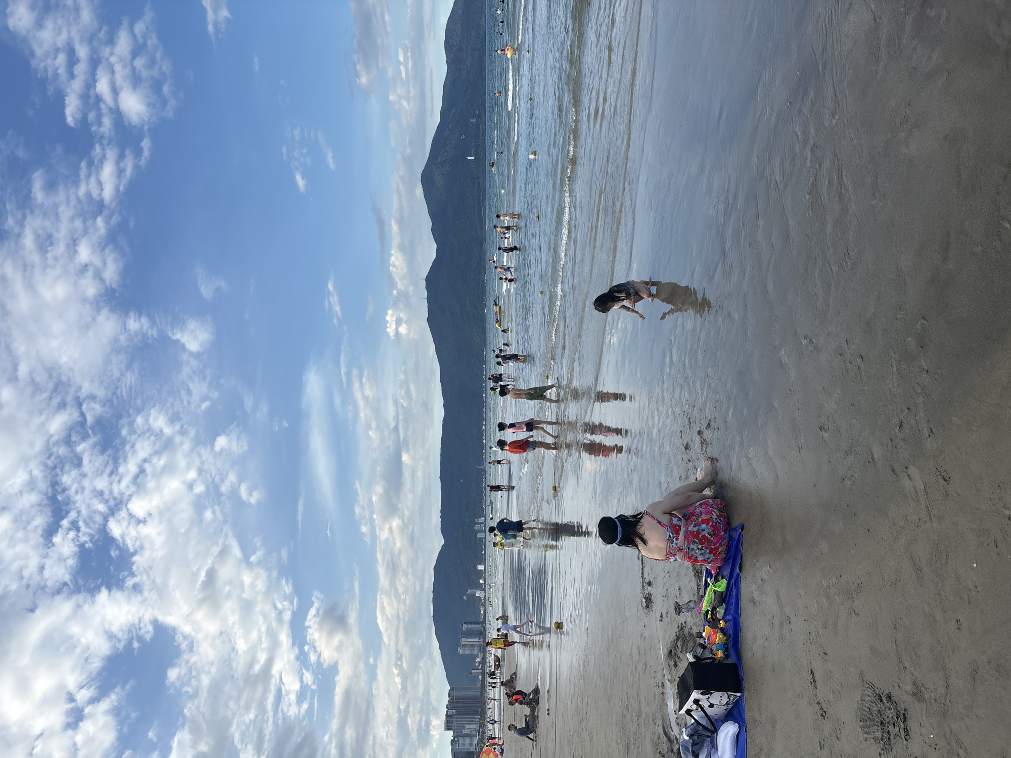 This is a vertical, eye-level, medium-shot photograph of a beach scene in Da Nang, Vietnam, likely in the vicinity of My Khe Beach, given the mountainous backdrop and urban development visible along the coastline. The image captures a wide expanse of sandy beach meeting the ocean under a partly cloudy sky.
In the foreground, a woman with dark hair, wearing a floral-print one-piece swimsuit and a dark headband, is seated facing away from the camera. She is positioned on a blue towel on the wet sand, with a black cooler bag and various children's beach toys, including sand molds and a shovel, arranged beside her. Her legs are extended towards the water, with her feet submerged in the shallow waves.
Further into the frame, closer to the water's edge, a young child with dark hair, wearing a light-colored outfit, is crouched down and appears to be interacting with the sand or water. This child is also facing away from the camera, and their reflection is visible in the wet sand.
Numerous other people are scattered across the beach and in the shallow water. Several children and adults are walking in the water, some appear to be playing or wading. In the mid-ground, there are clusters of people, and further out in the ocean, more individuals are visible, some swimming and others on what appear to be small watercraft.
The background features a long, undulating mountain range that forms the horizon line, topped by lush greenery. Along the base of the mountains, a dense urban landscape of high-rise buildings is visible, indicating proximity to the city. The sea is a moderate blue, with gentle waves rolling towards the shore. The sky is a vibrant blue with scattered white, puffy clouds that cast subtle shadows on the water. The overall lighting suggests it is late afternoon or early evening, with soft, diffused light. The sand itself is a wet, reflective surface in the foreground, showing ripples and tidal marks.
