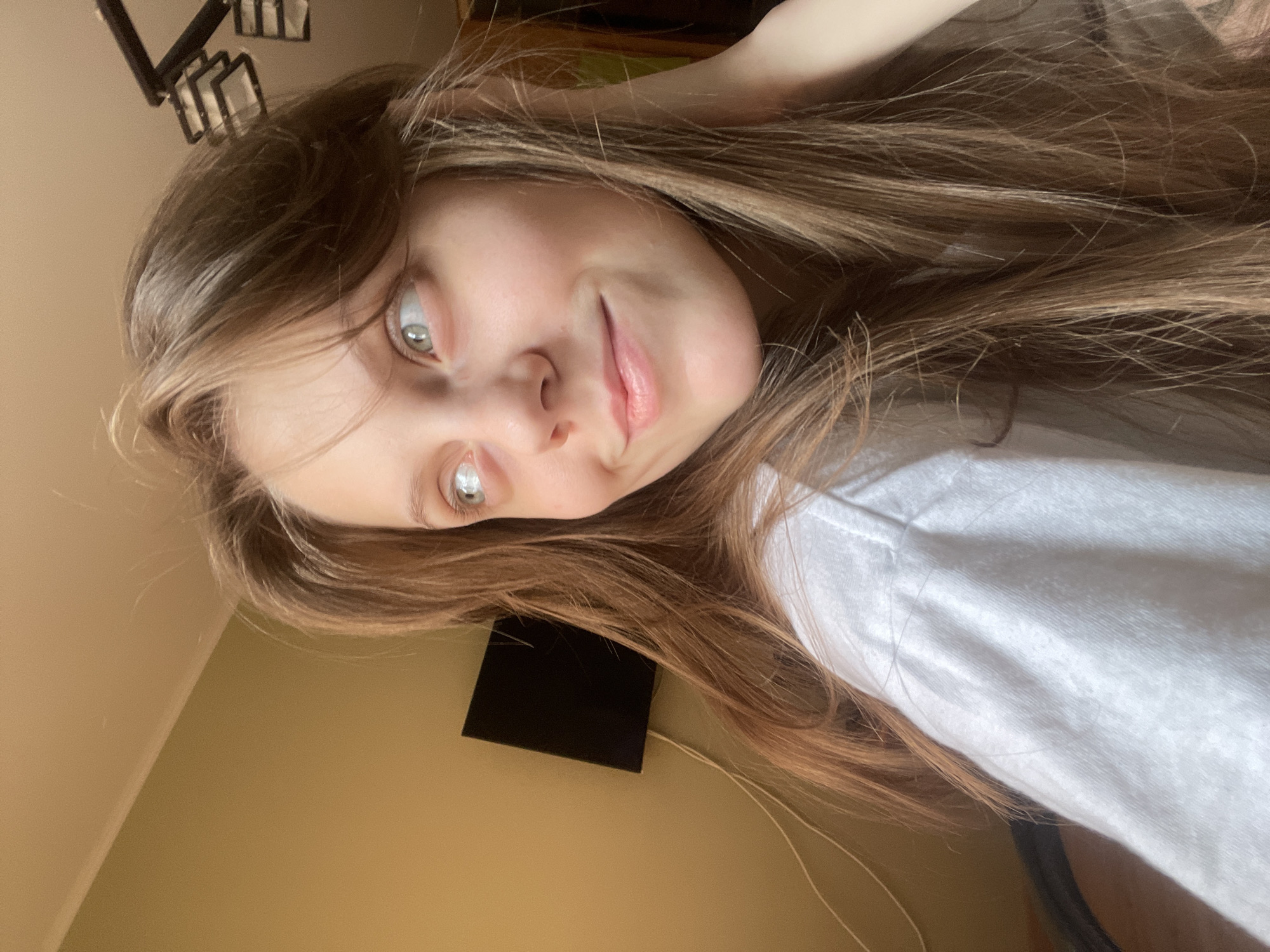 The image shows a close-up, chest-up shot of a young woman with long, light brown hair. She is wearing a white t-shirt and her head is tilted to her right, with her eyes looking upwards and to the right. She has a slight smile on her face and her right hand is touching her neck. The background is out of focus, but appears to be an indoor setting with a yellowish wall and a black television mounted on it. A light fixture is visible in the upper right corner. The image does not provide any specific information about the location in Ufa, Russia.