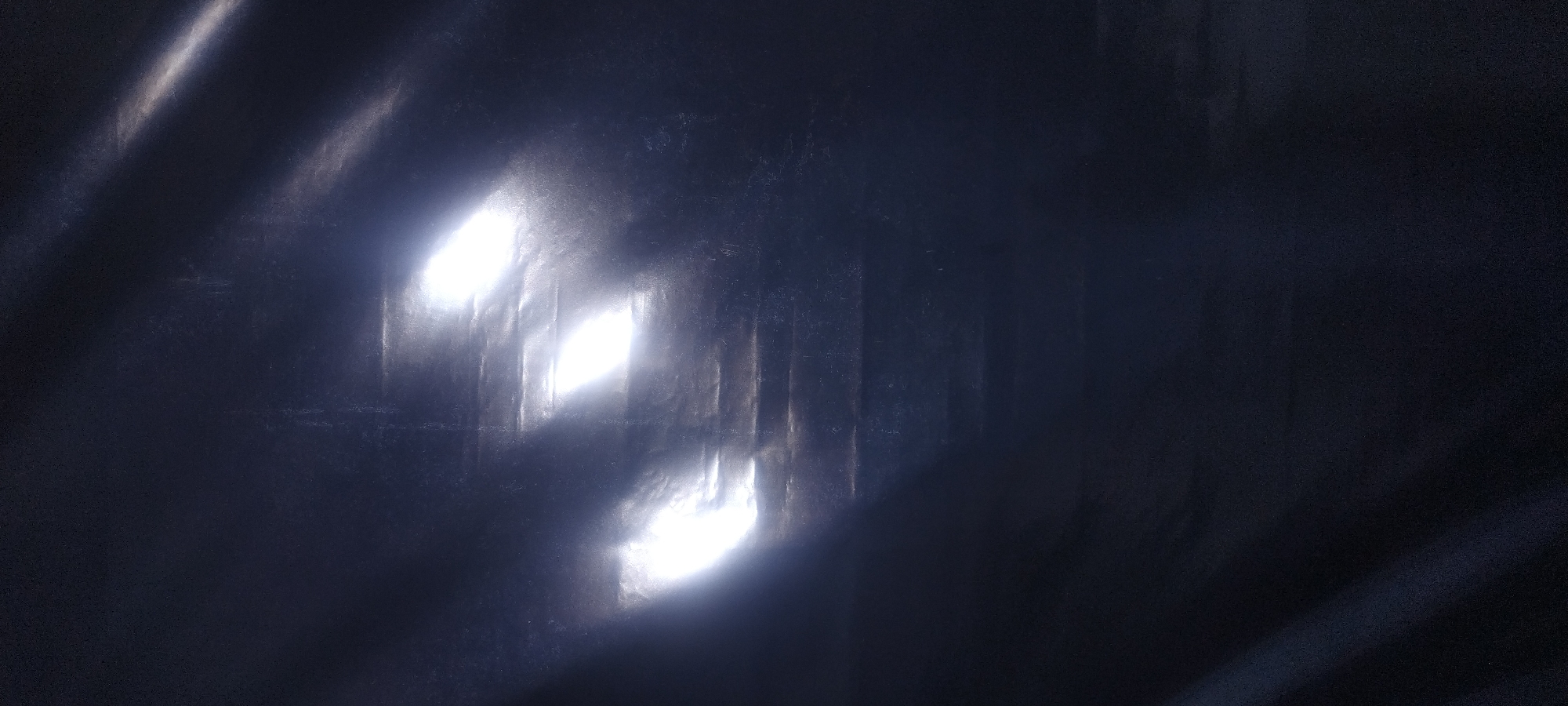 The image shows a dark, abstract scene with three bright, elongated light sources that appear to be reflecting off a dark, textured surface. The lights are arranged diagonally from the bottom left to the top right of the frame. The surface itself has a textured, somewhat crumpled appearance, with horizontal lines suggesting ridges or folds. There are also faint, lighter streaks or reflections across the surface, indicating the movement of light or a glossy material. The overall impression is one of dramatic lighting against a dark, possibly metallic or synthetic, background. There are no identifiable people, objects, or location details in the image.