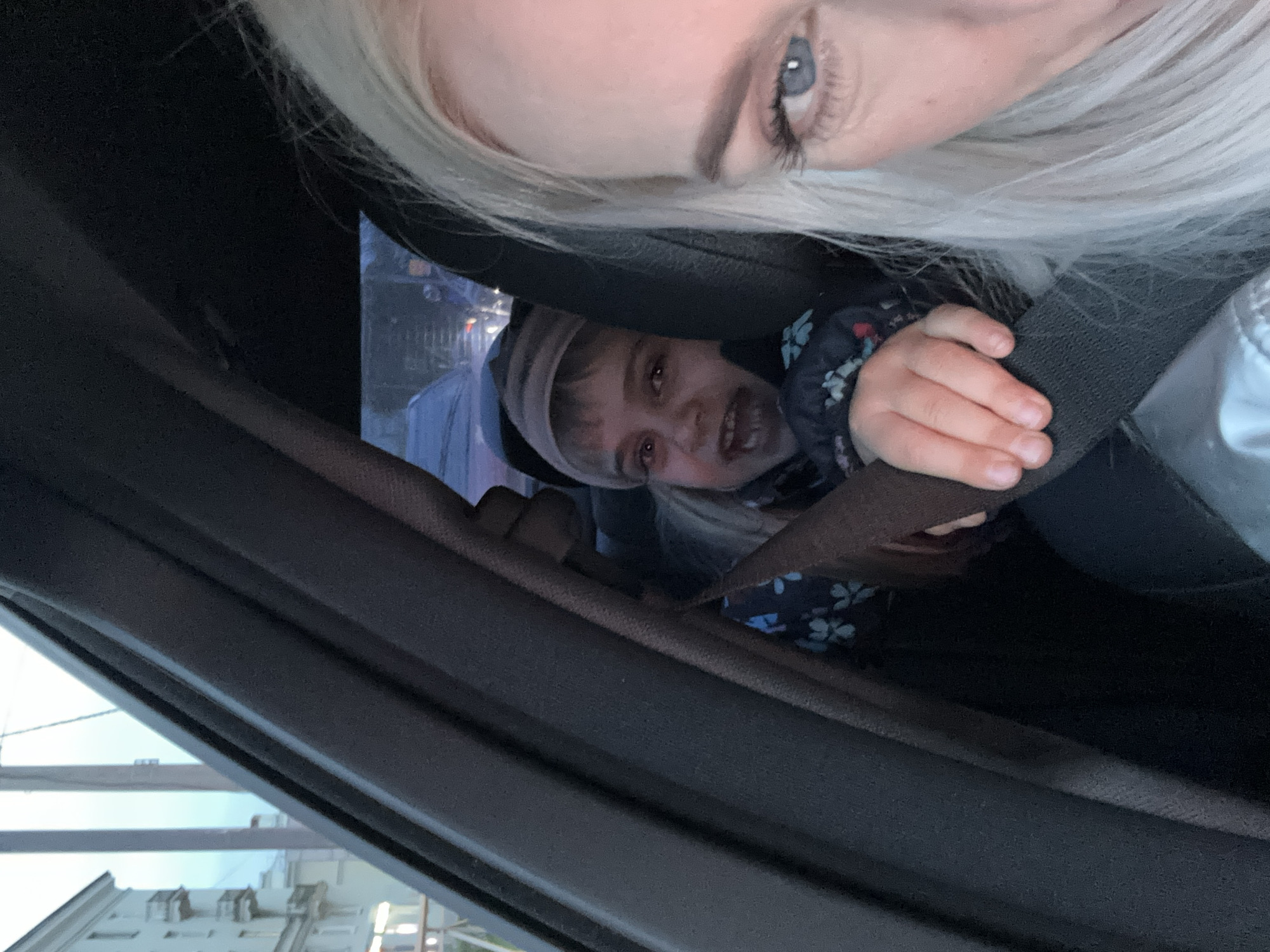 A selfie taken inside a vehicle captures a woman with blonde hair, partially visible in the top right corner, and a young child. The child, wearing a blue floral jacket and a light-colored hat, is secured in a seatbelt. The interior of the vehicle is predominantly dark. Through the window, the exterior environment reveals a building with multiple windows, suggesting an urban setting. Based on the architectural style of the building visible through the window, it can be inferred that the location is likely in Saint Petersburg, Russia.