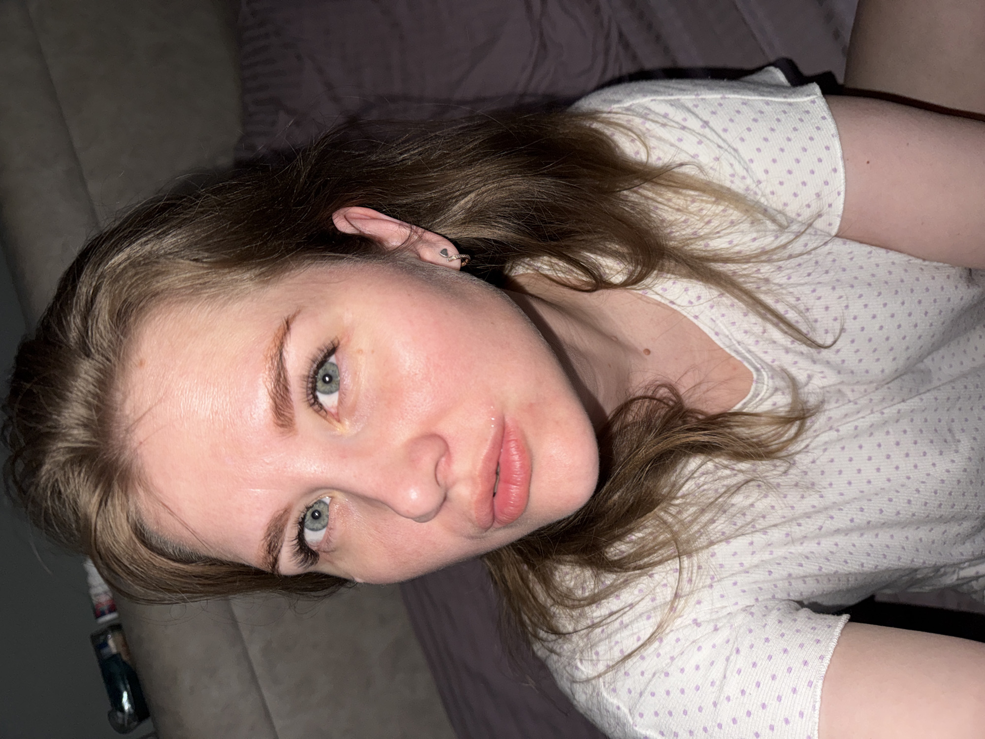 A pale-skinned, blue-eyed woman with brown hair is lying on a grey couch. She is wearing a white t-shirt with small lilac polka dots. Her hair is tousled around her head and shoulders. A silver earring is visible in her left ear. The scene appears to be indoors. The location is Gornyi (Dvurechenskoe S/P), Russia.