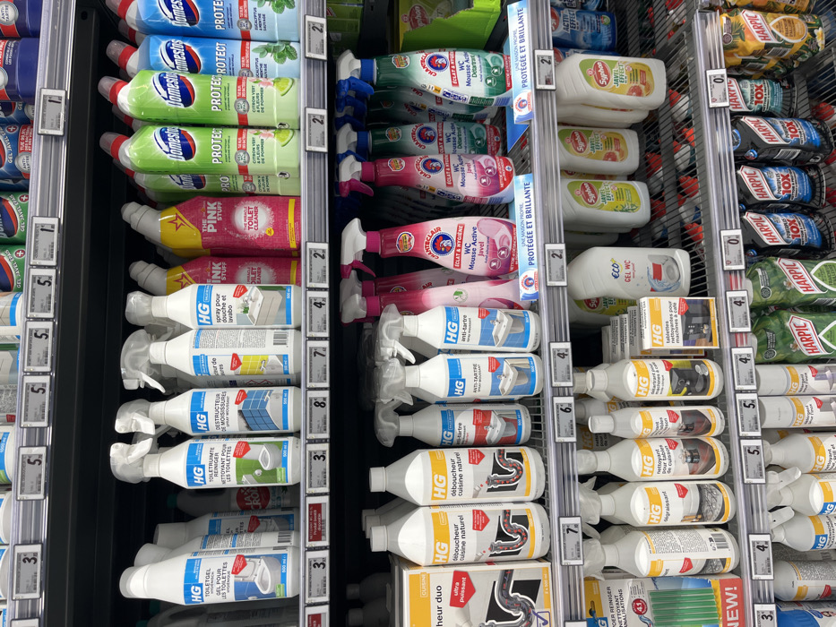 The image shows a grocery store aisle in Saint-Brice-Courcelles, France, at the coordinates 49.283, 3.999. The aisle is stocked with various cleaning products. Most of the products are in spray bottles with various colors, sizes, and labels. The aisle is organized by brands, and the top shelves are stocked with bleach-based products like Domestos. The middle shelves have various HG cleaning products for bathroom and kitchen use, while the bottom shelves are filled with Harpic toilet cleaner and other products. There is a price tag for each product, and the aisle has metal shelves with black borders. The aisle is lit by fluorescent lighting. The background is blurred out of focus.
