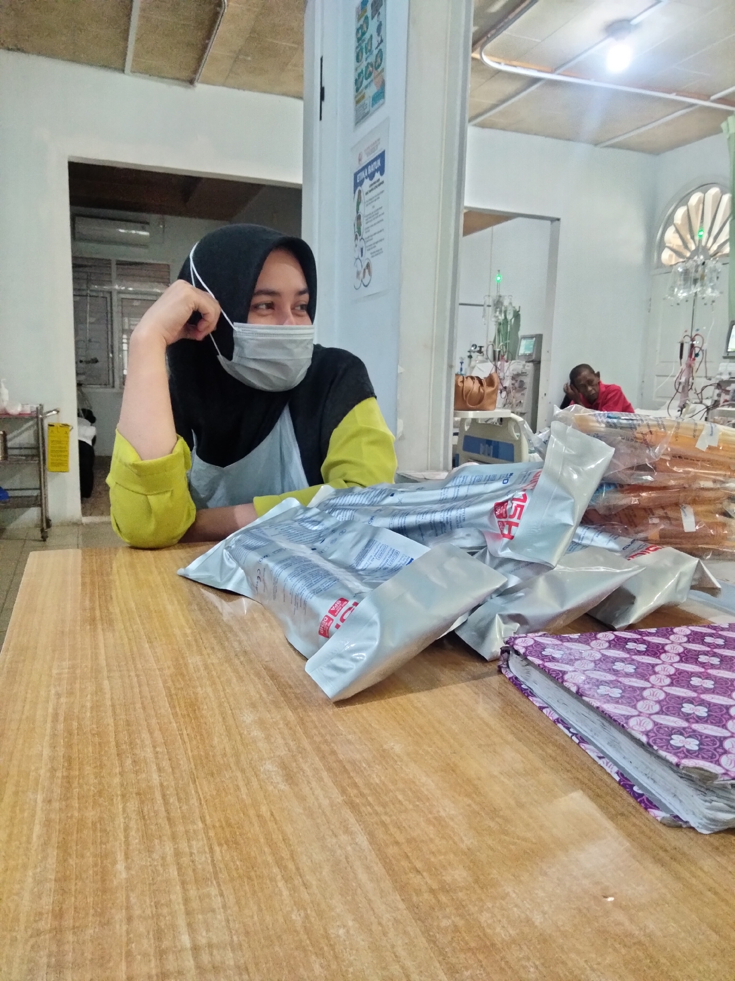 The image depicts an indoor medical facility, likely a clinic or hospital ward, in Lhokseumawe, Indonesia. In the foreground, a light-toned wooden table fills the lower half of the frame. Seated at the table on the left is a female individual wearing a black hijab, a gray protective face mask, a black long-sleeved shirt with bright yellow-green cuffs, and a light gray apron or panel over her torso. She rests her left arm on the table, hand near her face, and gazes towards the right side of the frame.
Piled on the table in the mid-ground are numerous silver foil-wrapped packages, likely medical supplies, with some visible red lettering that appears to be "FRESENIUS" or related branding. To the lower right, a purple-patterned book or folder with visible white pages lies partially on the table.
In the background, the room features white walls and a white ceiling with a single exposed light bulb. To the far left, a metal shelving unit with various items is visible. Through an open doorway on the left, an air conditioning unit is visible in an adjacent room. In the central background, a male patient with dark skin, wearing a red shirt, is visible reclining in a medical bed, resting his head on his hand. Behind him, medical equipment including IV poles with hanging bags, monitors, and potentially dialysis machines are present. A brown handbag rests on a nearby medical bed. On the right wall, a poster displaying text, including "ETIKA BATUK" (Cough Etiquette in Indonesian), is affixed. An arched window with vertical slats is prominently visible on the far right wall.
