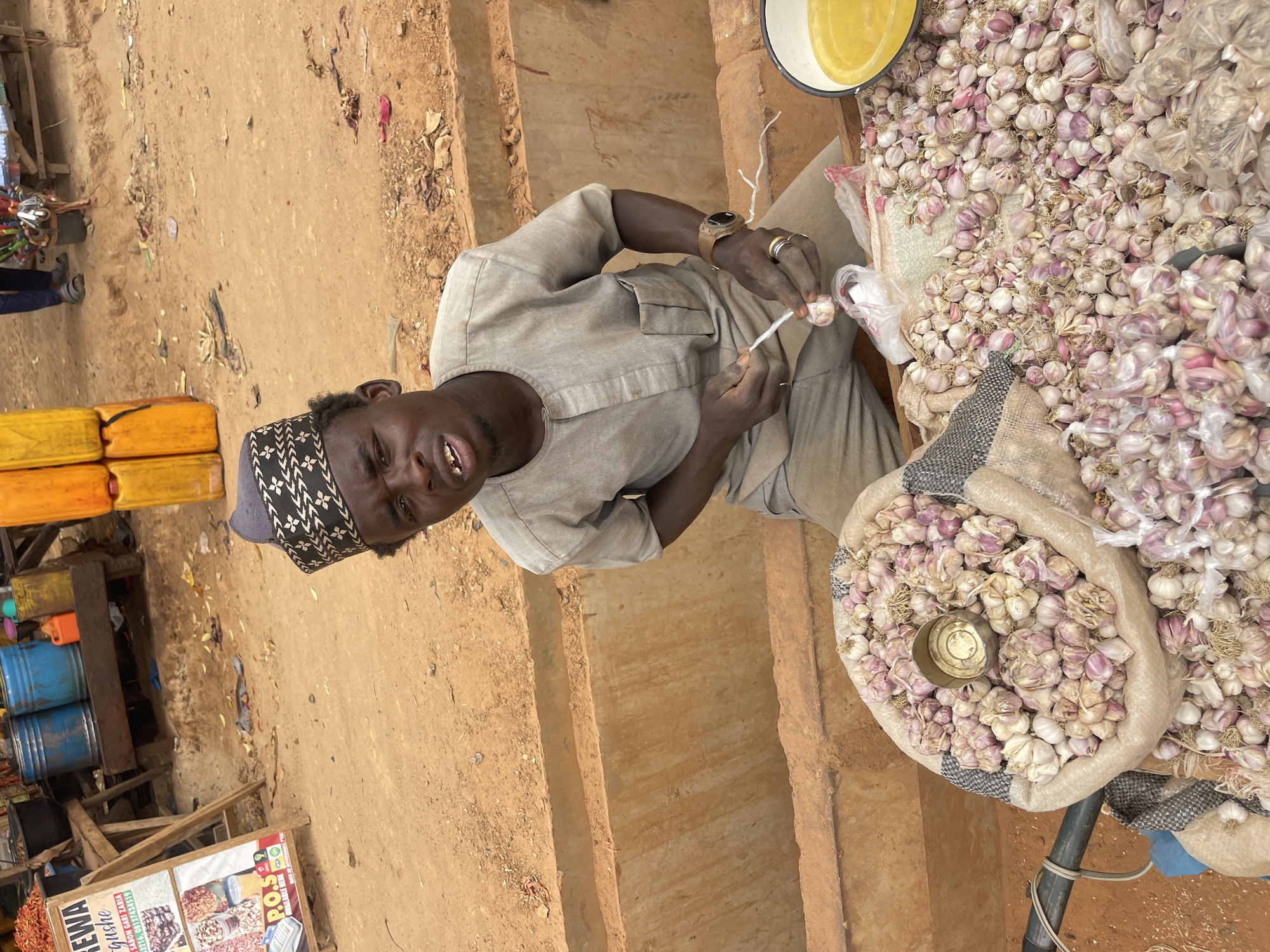 The image depicts a man in Zaria, Nigeria, engaged in selling garlic at an outdoor market. He is tying together garlic bulbs with white string. The man is dark-skinned, wearing a light grey traditional outfit and a patterned grey hat. He has a watch on his left wrist and rings on his fingers. Several bags and piles of garlic are visible, one containing a small metallic cup. To the left, there are stacks of yellow and blue containers, as well as a sign with food products and phone numbers. The ground appears to be made of reddish-brown soil. The setting is a marketplace with simple construction and a rustic feel.