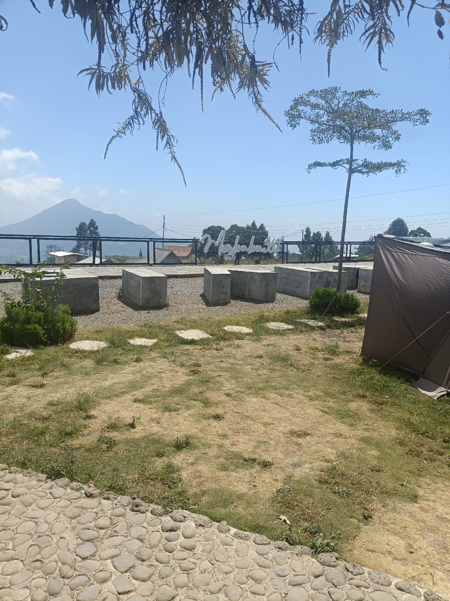 The image captures an outdoor seating area in Kopeng, Indonesia, with a scenic mountain view. In the foreground, a cobblestone path leads to a patch of dry grass and some scattered green vegetation. Several large, gray concrete blocks are arranged as seating in the middle ground, with gravel scattered around them. To the right, a brown tent is partially visible. In the background, a black metal fence separates the seating area from a view of houses and trees, with a prominent mountain in the distance under a clear blue sky. A tall, slender tree stands in the middle ground, and tree branches hang from the top of the frame. A white sign with text appears in the background.