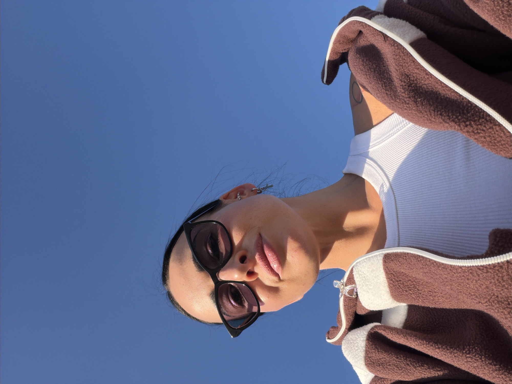 The image presents a low-angle portrait of a person set against a clear, bright blue sky. The subject, likely female, occupies the upper right portion of the frame, looking downwards towards the camera. They are wearing black cat-eye sunglasses, which reflect the sky and obscure their eyes. Their lips are slightly parted, revealing a subtle pink hue.
The person is dressed in a brown fleece jacket with distinctive white stripes or trim along the seams, worn over a white ribbed tank top. Accessories include multiple silver earrings in their left ear, with one featuring a small charm or chain. Dark hair frames the face, pulled back from the forehead.
The background is a vast, uninterrupted expanse of vibrant blue sky, suggesting an outdoor setting on a sunny day. Strong, direct sunlight is evident from the distinct shadows cast on the person's neck and jawline. No other objects, landscapes, or structures are visible. The image captures a casual moment with a focus on the subject against a minimalist, bright backdrop. There is no visible text in the image.