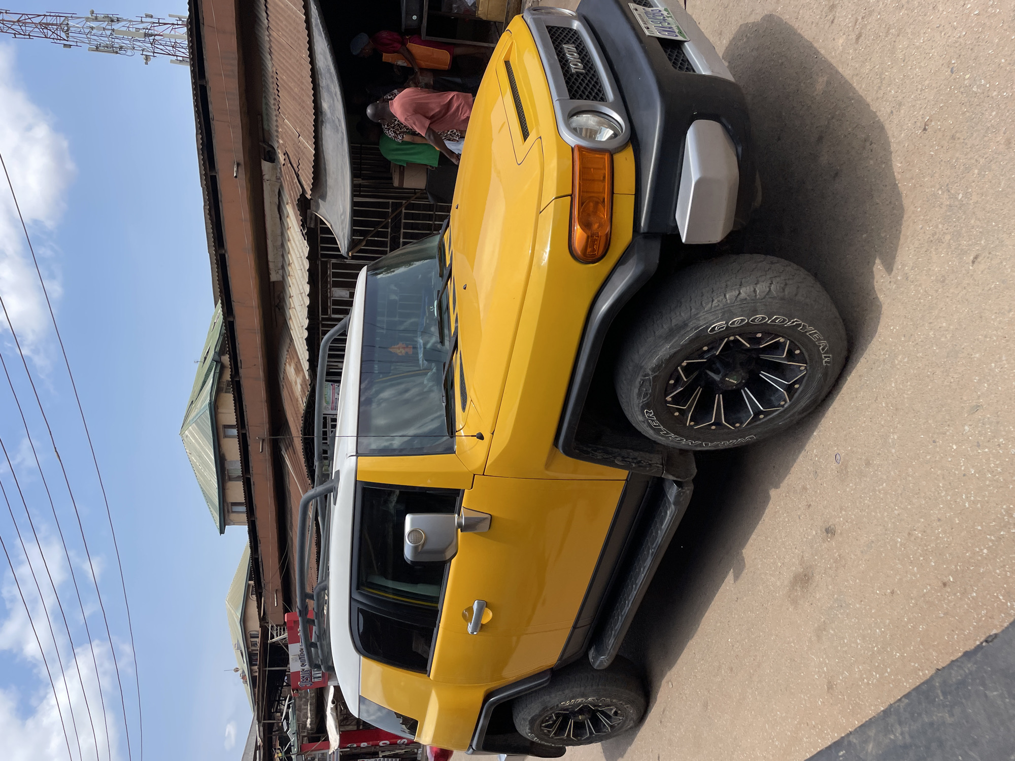 The image displays a bright yellow and white Toyota FJ Cruiser SUV, primarily viewed from its front-right side, on a sunny day in Onitsha, Nigeria. The vehicle is either parked or moving slowly on a paved surface, casting a clear shadow. The front grille features the "TOYOTA" emblem, and the visible license plate reads "GMN-987XQ", indicative of a Nigerian registration. The tires are branded "GOODYEAR WRANGLER".
In the background, a street or market scene unfolds, characterized by buildings with weathered, corrugated metal roofs. Blurry figures of people can be discerned near these structures, suggesting ongoing daily activities. The sky is largely clear with a few faint clouds, and power lines traverse the upper left portion of the frame. A large telecom tower is also visible in the distance, further signifying an urban setting. The strong daylight and distinct shadows confirm it is daytime.