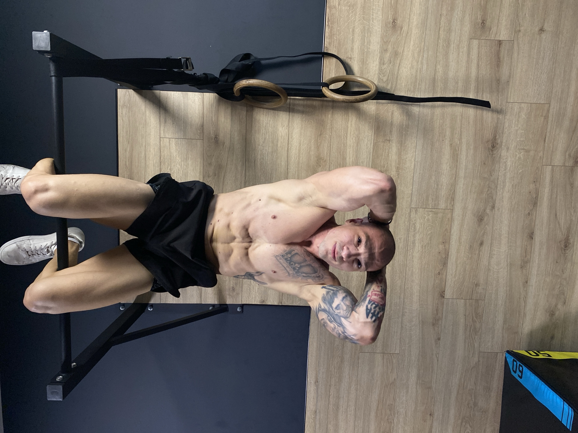 A muscular man with tattoos is lying on his back on a weight-lifting bench in what appears to be a gym. He's wearing black shorts and white shoes. His hands are positioned behind his head. Above him, gymnastic rings are hanging from the pull-up bar. The gym has a light wood-grain floor, and the walls are a combination of black and white. It's not possible to verify the location within Pleven, Bulgaria, based on this image.