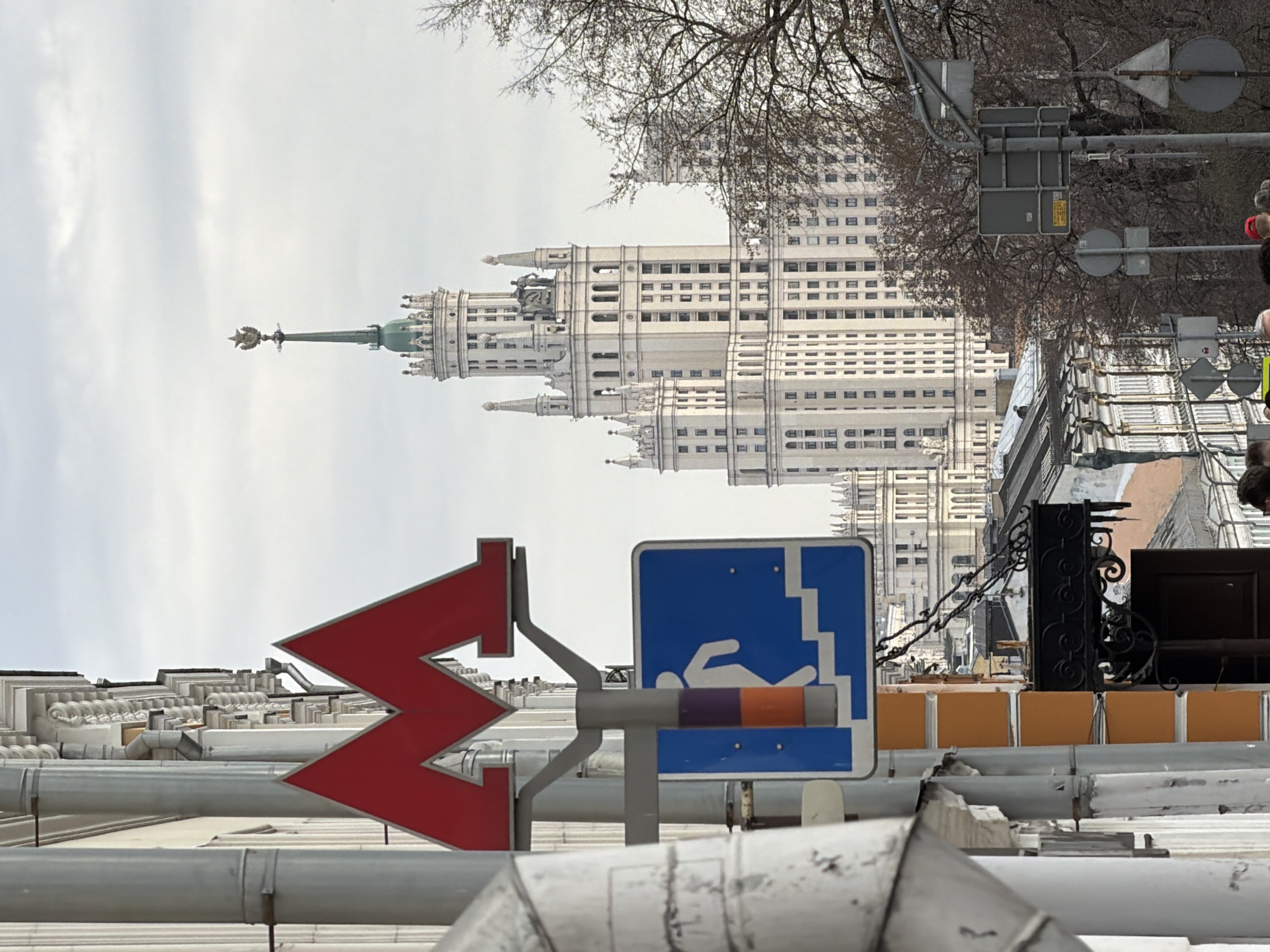 The image, taken in Moscow, Russia, depicts a view of one of the Seven Sisters skyscrapers, specifically the Kotelnicheskaya Embankment Building, towering over a city street. In the foreground, a red Moscow Metro sign (a stylized "M") and a blue sign indicating an underground pedestrian crossing are prominently displayed. The signs are attached to metal poles and are partially obscured by the city's infrastructure, including pipes and rooftops. Bare trees and traffic signals are visible near the building. The sky is overcast.