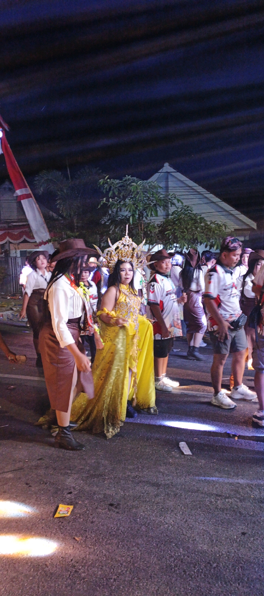 This image captures a nighttime scene in the city of Kasiyan, Indonesia, likely during a parade or cultural event. The foreground features a group of individuals, with a woman in a striking, ornate golden costume and elaborate headpiece at the center. She is holding a yellow object in her hand. To her left, a person wearing a brown cowboy hat and a white shirt with brown details is visible. Other individuals in white shirts with red and black accents, and brown cowboy hats, are also present, some holding what appear to be flags or banners. The background reveals trees, a building with a distinctive triangular roof, and a red and white flag, possibly the Indonesian flag, flying from a pole. The ground is asphalt, illuminated by stage lights creating bright spots. A small yellow packet with the number "2" printed on it is visible on the road. The overall atmosphere suggests a lively and festive event taking place outdoors after dark.