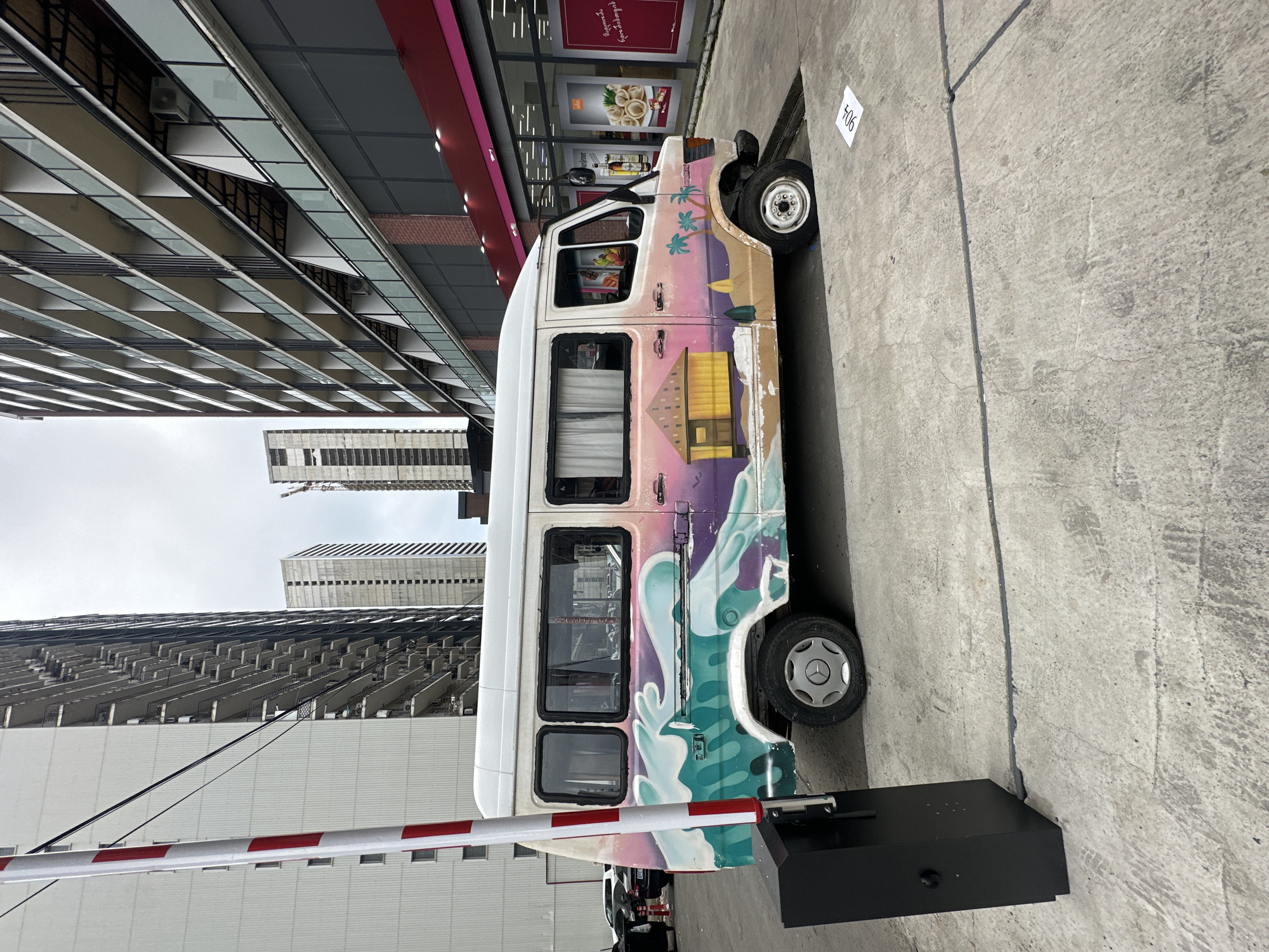 A white van with colorful artwork is parked on a concrete surface in front of a modern building complex. The van's artwork depicts a beach scene with waves, palm trees, and a hut. The building in the background features multiple floors with balconies and large windows. A red and white striped pole and a black barrier are to the left of the van, suggesting an entrance or exit. The scene appears to be in an urban environment, possibly a street or a parking area in Batumi, Georgia, given the architectural style of the buildings. The lighting suggests it is daytime, with overcast skies. There are no people visible in the frame.