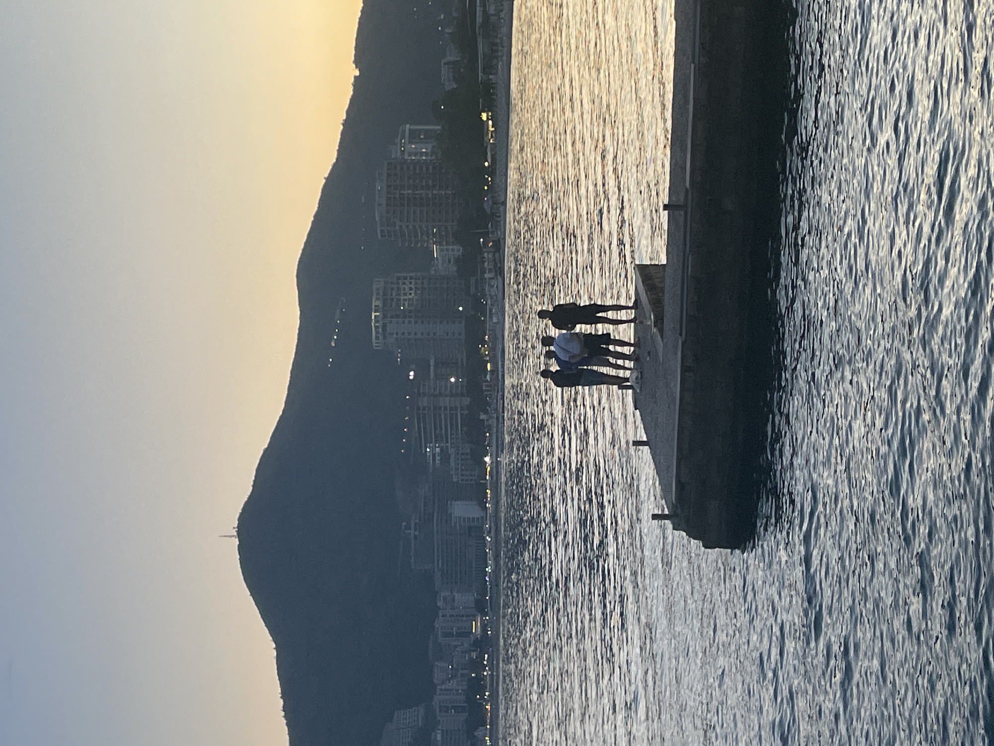 A sideways view of the Adriatic Sea, Budva, Montenegro at dusk. From left to right: the skyline is dominated by a large mountain with lights from the city extending from the base to the top. In the center, the water of the sea reflects the sky and city lights. On the right, a concrete pier extends into the water, and four people are standing at the end of the pier.