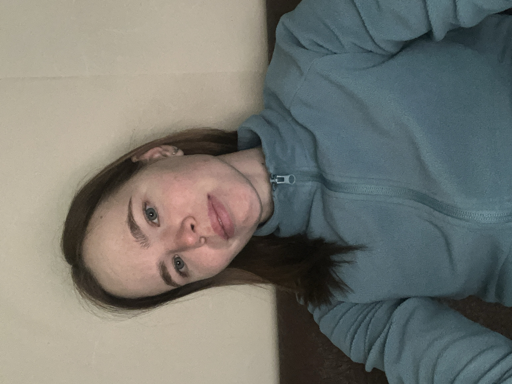 A woman with brown hair and blue eyes is wearing a blue fleece jacket with a zipper. She appears to be indoors, with a light beige wall visible in the background. The photo does not contain sufficient information to determine her location in Chaikovsky, Russia or any other specific location, action, interaction, or relevant objects.