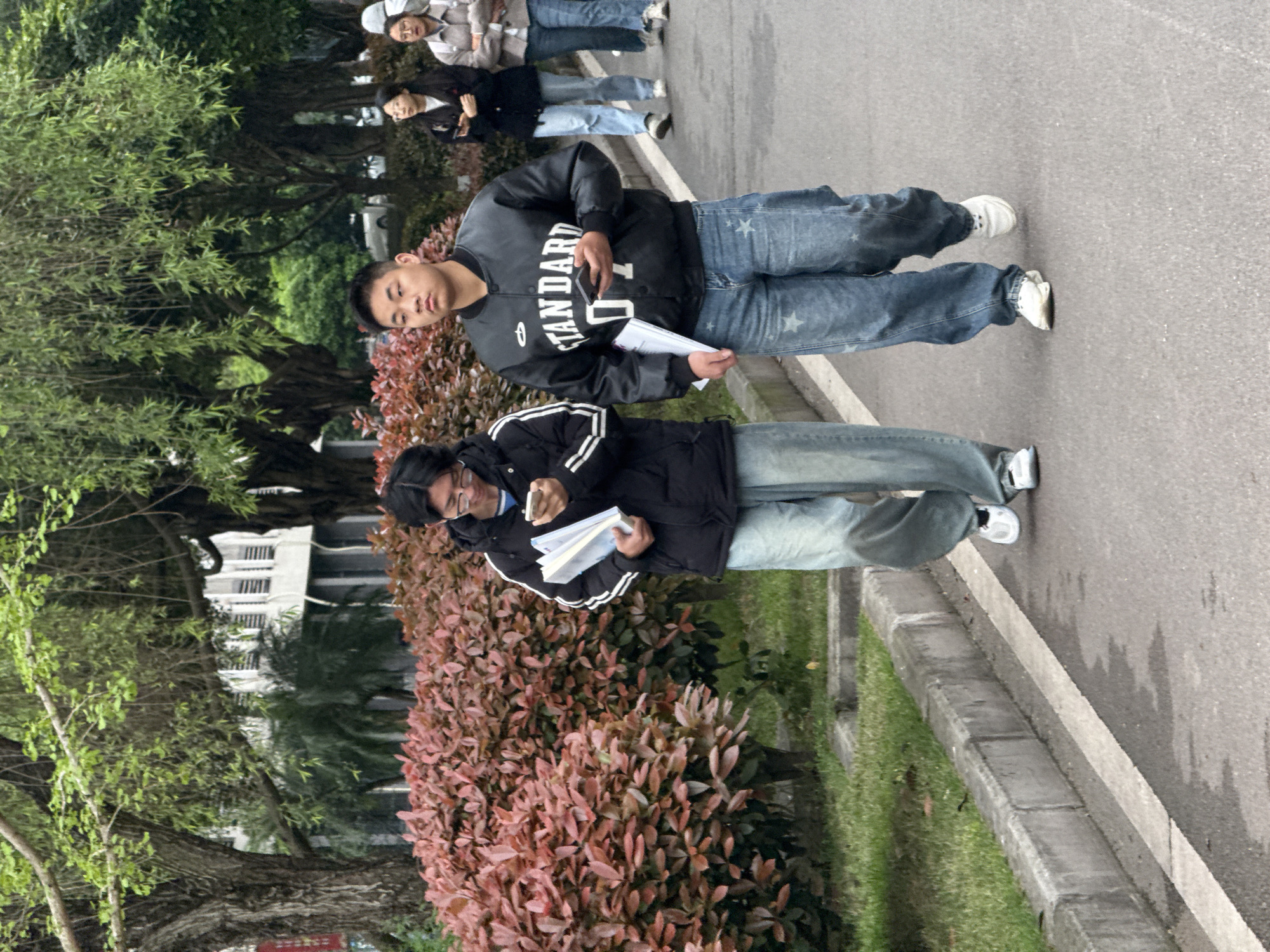 Two young men are standing on a paved path. The first man is wearing a black jacket with "STANDARD" printed on it, blue jeans with white stars, and white sneakers. He holds a smartphone in one hand and papers in the other. The second man wears a black jacket with white stripes, blue jeans, and white sneakers, holding papers in one hand and a phone in the other. A group of people walks behind them. The paved path is next to a strip of grass and a hedge with reddish leaves. Trees and a building are visible in the background. This scene appears to be in Jiangjin, China.