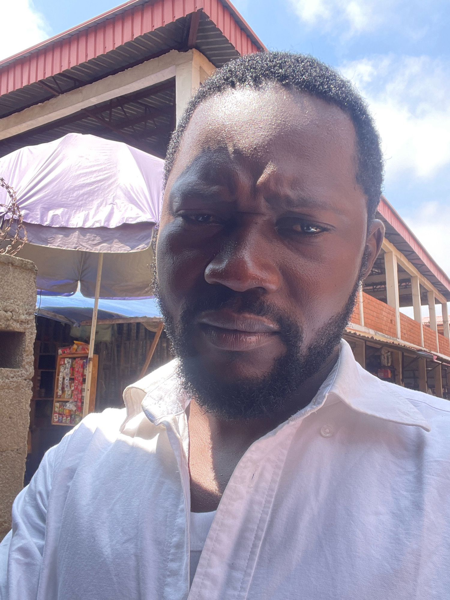 A close-up selfie shows a Black man with dark skin and a beard, wearing a white collared shirt over a white undershirt. He has short, dark, curly hair and is looking directly at the camera with a furrowed brow. The background depicts an outdoor marketplace with a red corrugated metal roof extending from the top of the frame. To the left, a purple tarp is stretched over a stall, and behind the man, a two-story building with a red brick lower level and concrete upper level is visible. The environment suggests a sunny day, with some clouds in the sky. The scene is likely in the city of Kubwa, Nigeria, given the architectural style and market setting.