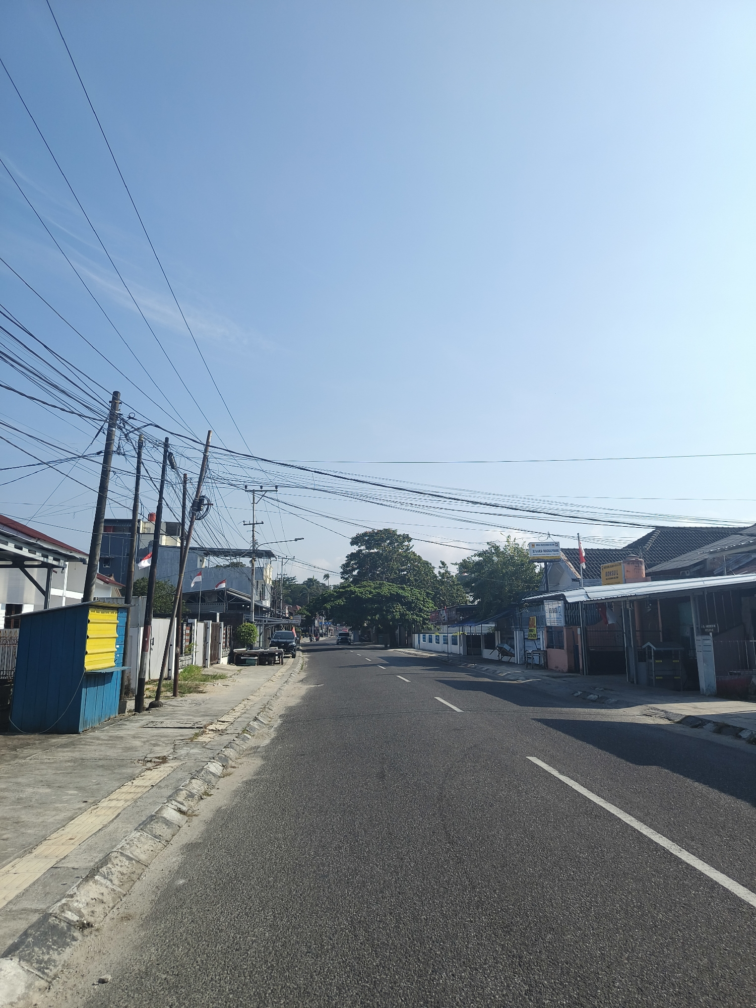 The image depicts a street scene in Pangkalpinang, Indonesia, characterized by a clear blue sky and overhead power lines. The road, paved with asphalt and marked with a white line, stretches into the distance. On the left side of the road, a sidewalk is visible, bordered by a low concrete curb. Adjacent to the sidewalk is a structure with yellow and blue panels, and behind it, a building with white walls and a gray roof. Further down the street on the left, several other buildings with varying architectural styles and roof types are present.
The right side of the road features a sidewalk and a series of commercial buildings and structures with tiled roofs. Signs are visible on some of these buildings, including one that reads "KONSUL." A few vehicles, including a black car and a trailer, are parked or moving along the road. Lush green trees are interspersed with the buildings, contributing to the urban landscape. The overall impression is of a typical street in a built-up area of Pangkalpinang, likely during daylight hours given the bright sky and shadows. The scene appears calm and orderly, with no evident human activity beyond vehicles on the road.