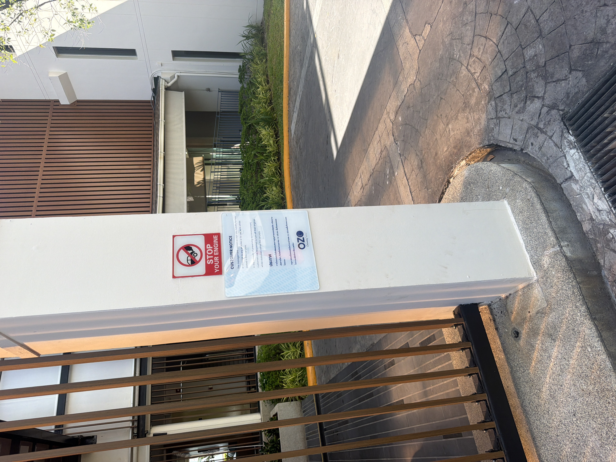 The image captures a close-up of a white concrete pillar located at what appears to be an entrance or gate, likely in Baan Chaweng, Thailand. Attached to the pillar is a red and white square sign with a circular icon depicting a car with a diagonal line through it, and the text "STOP YOUR ENGINE" below. Beneath this sign, a larger rectangular notice is affixed, containing text in both English and what appears to be Thai. The English text, partially visible, states "CUSTOMER NOTICE" and further text about the management not accepting liability for loss or damage to any vehicles or its contents parked in this car park. The notice also mentions "In the interests of preserving our environment, customers are requested to turn off their engines whilst stationary within this car park." The lower portion of this notice displays the "OZO" logo, followed by "Chewing Samui." Behind the pillar, vertical wooden slats forming a fence or gate are visible, along with a building with a modern architectural style featuring vertical wooden panels and white walls. The ground in front of the pillar is paved with decorative stone tiles, and a drain grate is present in the foreground. Lush green vegetation is visible in the background, suggesting an outdoor setting, possibly a hotel or resort entrance. The lighting indicates it is daytime, with sunlight casting shadows on the ground and highlighting the pillar.