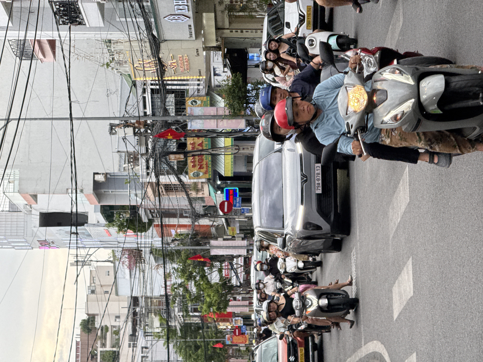 The image depicts a busy street scene in Nha Trang, Vietnam, likely during daylight hours. The foreground is dominated by a procession of motorcycles and scooters navigating traffic. Several individuals are visible on these vehicles, many wearing helmets and casual clothing. One scooter in the center foreground has a rider wearing a blue hooded jacket and patterned pants, with the motorcycle's headlight illuminated. Behind them, another rider is on a red and black scooter. Further back, a grey car with a license plate reading "79A-878.13" is prominent.
The background reveals a dense urban environment with multi-story buildings lining the street. Signs for businesses such as "SPA" and "XB HN" are visible on the storefronts. Overhead, a complex network of electrical wires crisscrosses the sky, a common sight in many Vietnamese cities. Red and yellow Vietnamese flags are displayed on poles along the street. The overall atmosphere suggests a bustling commercial area with active transportation and pedestrian activity.