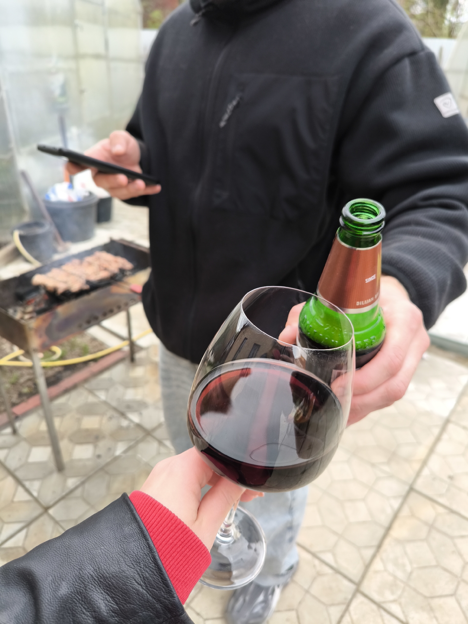 In an outdoor setting, a person holds a glass of red wine, toasting towards another person holding a green beer bottle and a smartphone. A grill with meat is in the background. The location is possibly in Novofryazino, Russia, based on the brand of beer. The ground has a tile pattern. The person holding the beer is wearing a black jacket and jeans. The person holding the wine is wearing a black jacket with a red sleeve.
