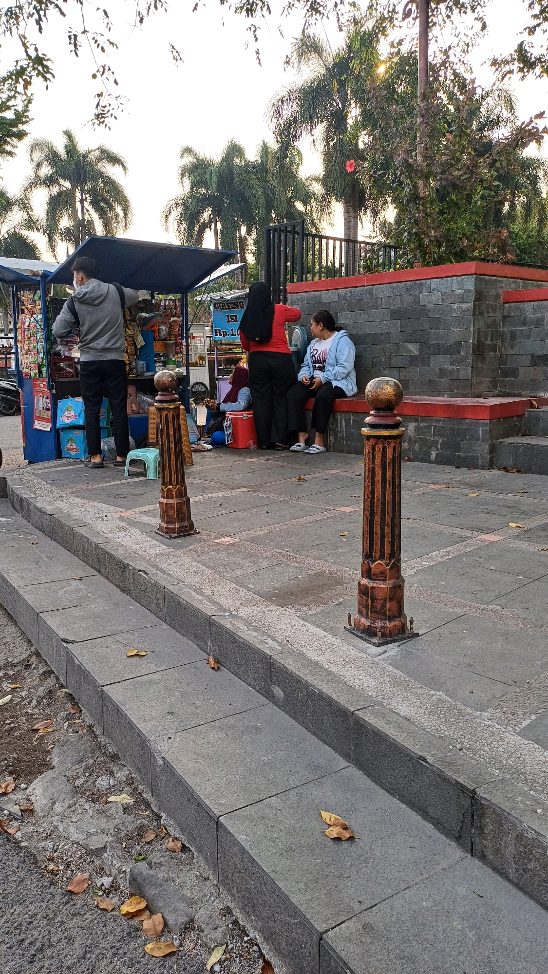 This image shows a street scene in Klaten, Indonesia. A vendor stall, likely selling snacks or small goods, is set up on the left side of the frame. A man wearing a grey hooded jacket and a face mask stands behind the stall, interacting with the vendor. Two women are seated on a red bench to the right of the stall. One woman is wearing a red top and black pants, and the other is wearing a blue hoodie and dark pants. Several palm trees are visible in the background, suggesting a tropical or subtropical environment. The ground is paved with stone tiles, and there are decorative posts with spherical tops lining the walkway. Fallen leaves are scattered on the ground and steps, indicating it might be autumn or a period of leaf fall. The overall scene depicts a casual outdoor setting in a public space.