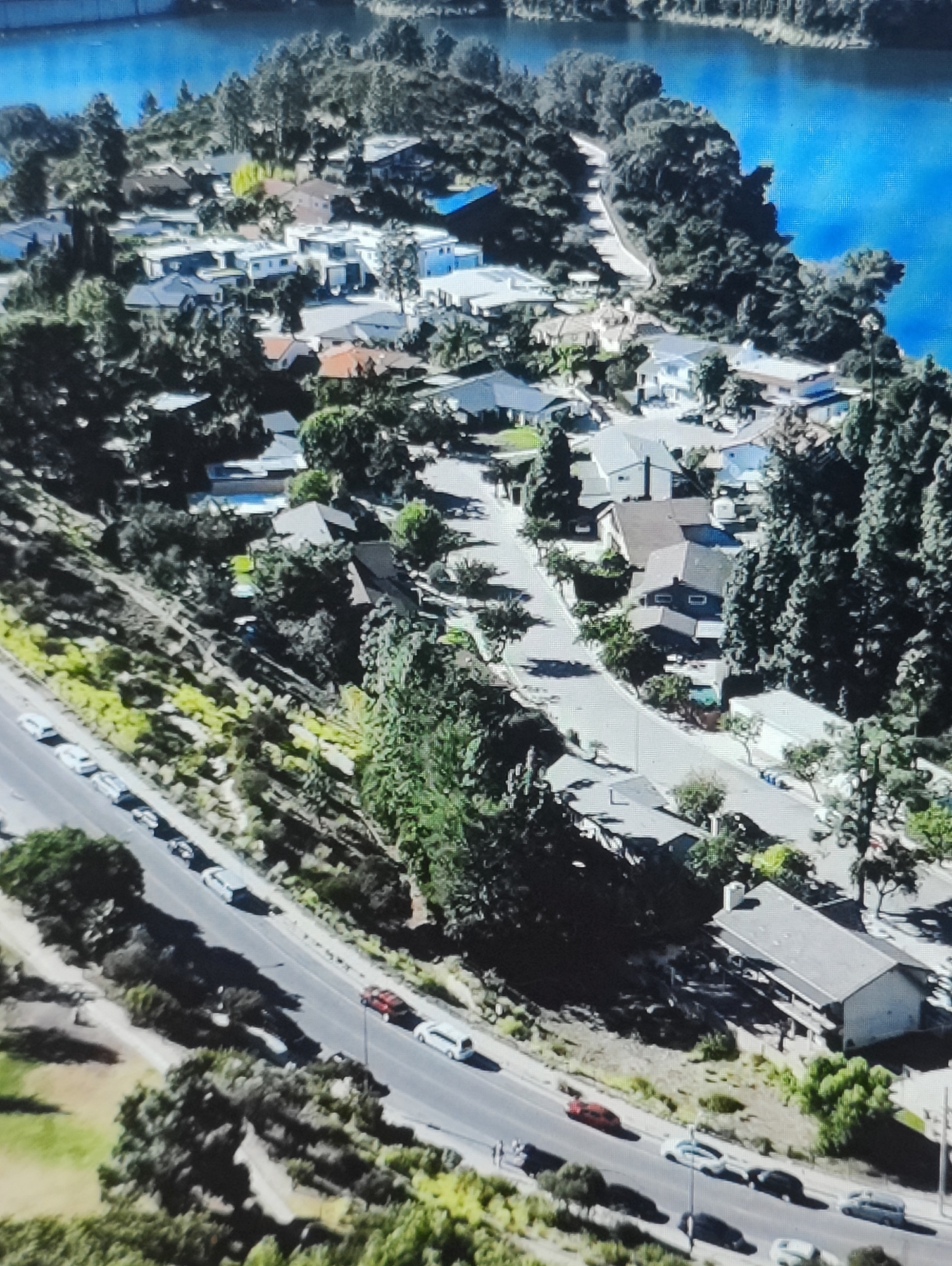 The image shows an aerial view of a suburban neighborhood situated on a hillside overlooking a body of water. The houses are of varying architectural styles, mostly single-family dwellings with pitched roofs. Many are surrounded by mature trees and landscaping. A road winds through the neighborhood, with several cars parked along its sides and one in motion. In the upper portion of the image, the blue water of a lake or reservoir is visible, bordered by dense vegetation and the distant shoreline. The scene suggests a residential area in a hilly, possibly coastal or lakeside, environment.