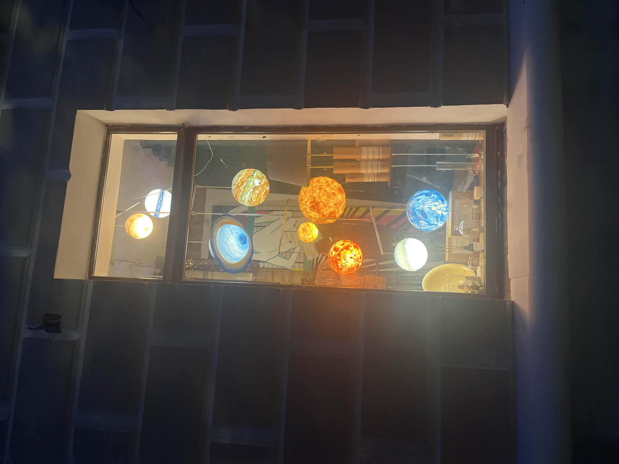 The media file depicts a dimly lit room with a window showcasing a display of illuminated spherical objects resembling planets. The window is framed by a textured wall, suggesting an indoor setting. The objects are varied in color and pattern, with some appearing fiery orange and yellow like the sun, while others are blue and white, reminiscent of Earth. They are suspended from wires, creating a solar system model.
Inside the window, beyond the planet display, are shelves filled with various items. These include what appear to be books, canisters, and other decorative objects. The lighting suggests it is nighttime or the room is intentionally kept dark, with the planets being the primary light source in the immediate view. The overall mood is somewhat mysterious and educational, possibly a display in a home, school, or science center. There are no people visible in the frame. No visible text is discernible from the image. The location context is Krasnodar, Russia.