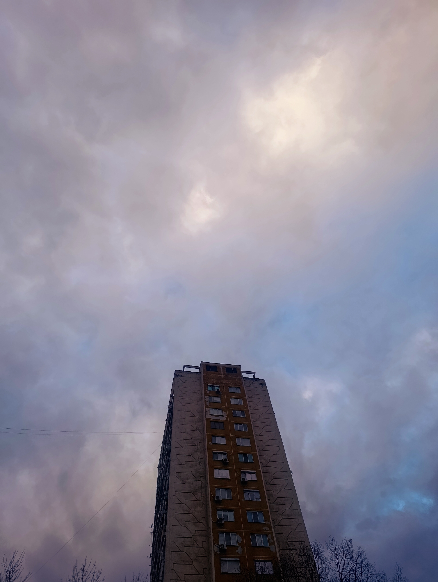 An upward angle shot depicts a tall building against a cloudy sky. The building, likely residential, features multiple rows of windows and a concrete facade that appears aged and somewhat weathered. Some windows have air conditioning units installed. Bare tree branches are visible at the bottom of the frame, suggesting an autumn or winter setting. The sky is filled with clouds, ranging in color from light grey to dark blue, with a patch of bright light filtering through the clouds above the building. The scene is likely located in an urban environment, possibly Kardzhali, Bulgaria, based on the prompt. No people are visible in the image.