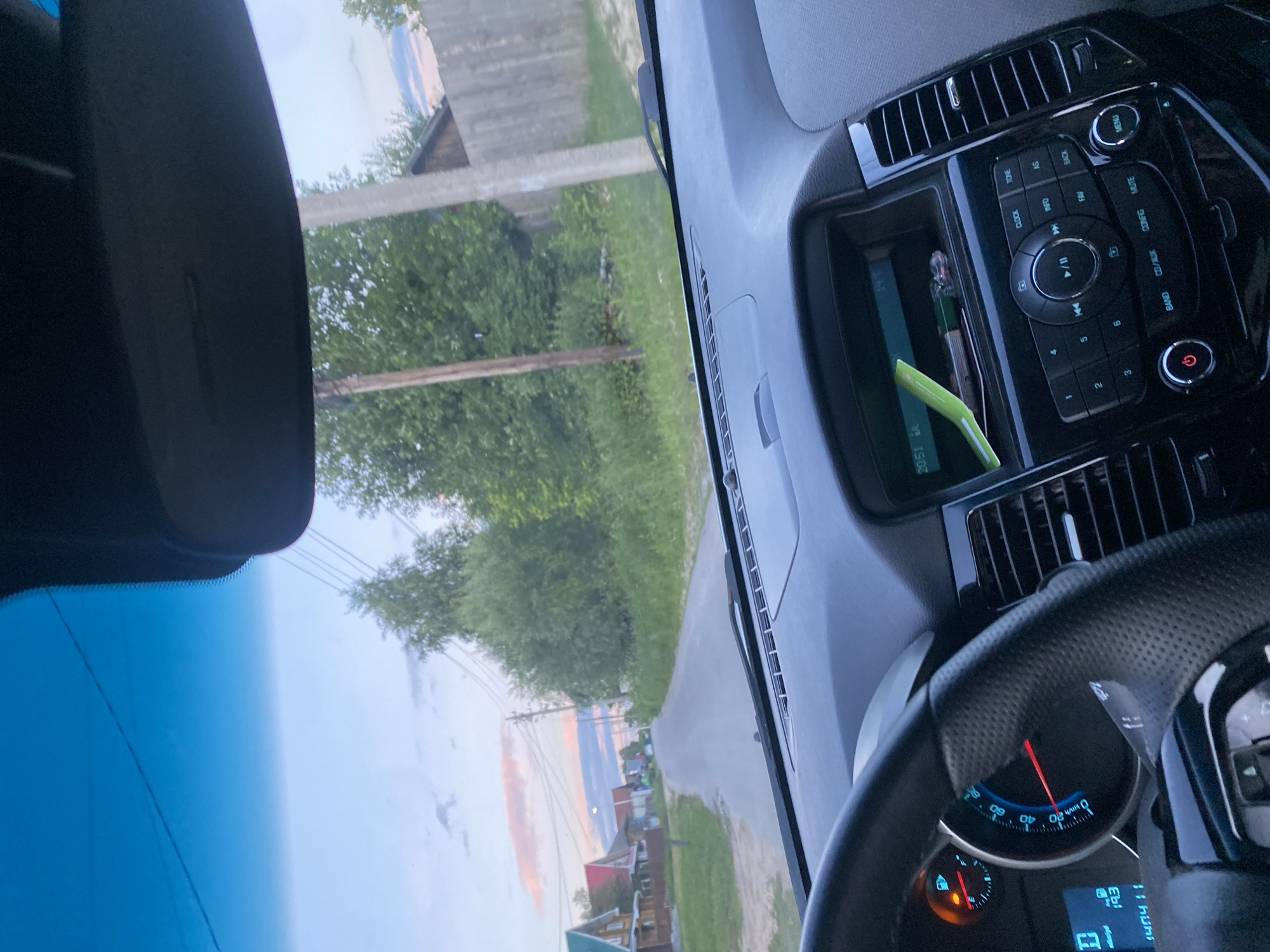 The image is a first-person view from inside a car, looking through the windshield. The car appears to be in motion on a paved road in a rural or suburban area, possibly Медведево, Russia, given the presence of wooden houses with colorful roofs, a wooden fence, and dense trees lining the road. The sky is partly cloudy with soft pink and blue hues, suggesting it is either sunrise or sunset. The car's dashboard is visible, including a steering wheel, speedometer showing approximately 0 km/h, a digital display with the date "14.07.19", and a car stereo with a green object on its screen. The overall scene depicts a peaceful drive through a quiet residential area during twilight.