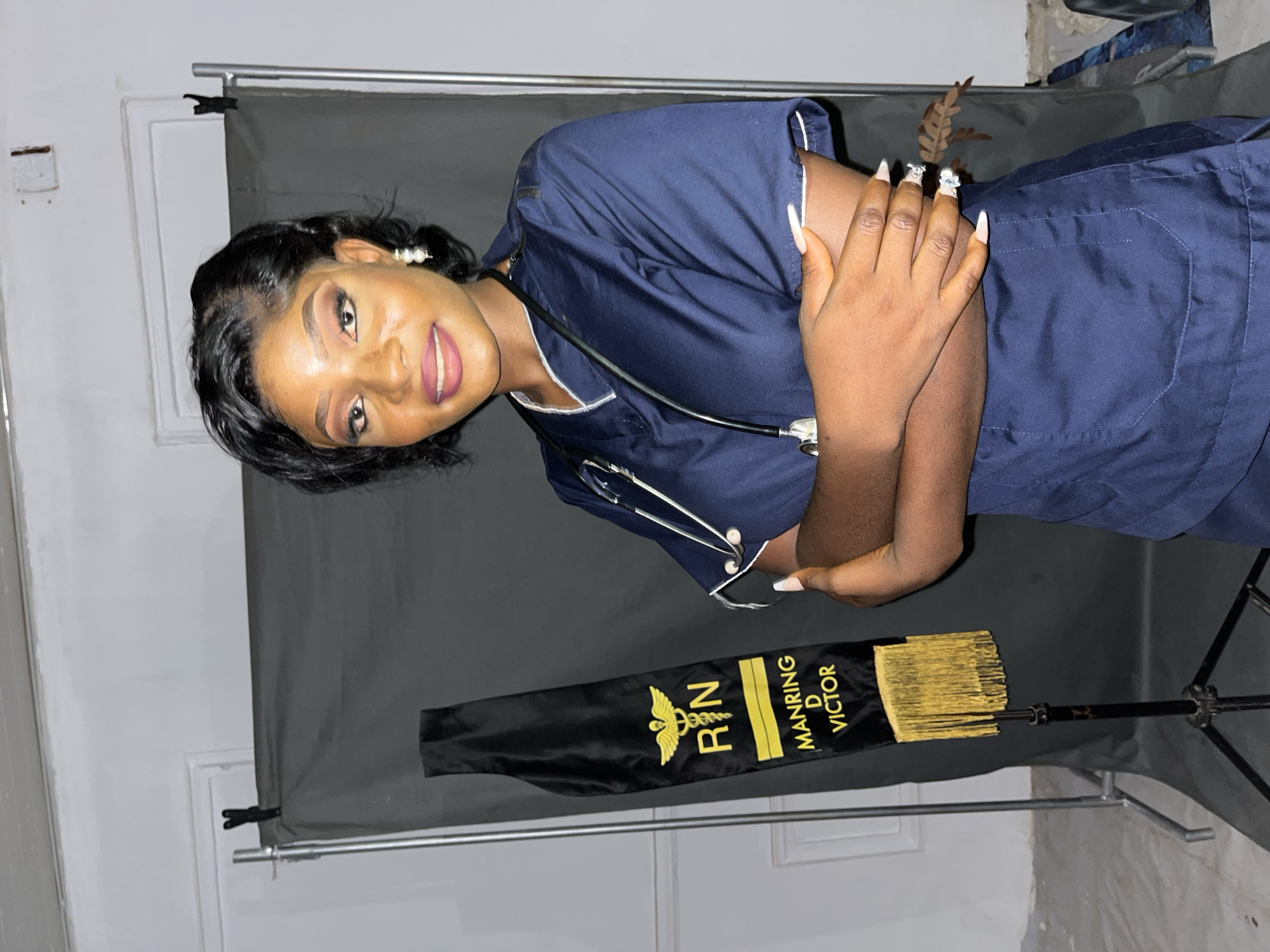 A medium shot shows a woman in a dark blue medical scrub suit. She is wearing a stethoscope around her neck. She has dark hair and long white nails. Her hands are clasped together. She is smiling. A black sash with the words "R N" and "MANRING D VICTOR" in gold lettering is in the foreground. A black photography backdrop is in the background. The location is Unknown, Unknown.