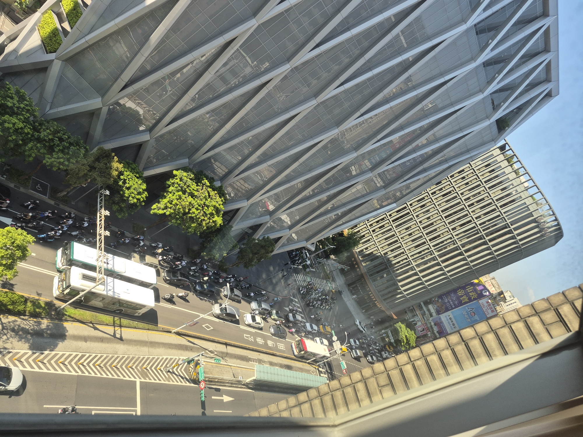 An aerial, high-angle, daylight shot captures a street scene in Taipei, Taiwan, characterized by modern architecture and urban traffic. In the foreground, a distinctive building features a facade of staggered, diamond-shaped panels with a metallic sheen, reflecting the bright sky. To its right, a taller, cylindrical building with a patterned concrete exterior curves along the street. The street itself is a busy artery, filled with a variety of vehicles including numerous motorcycles, cars, and two white buses traveling in the same direction. Pedestrians are visible on the sidewalks and crossing the street, some appearing to be part of a procession or gathering. Lush green trees are interspersed throughout the urban landscape, providing a natural contrast to the concrete and glass structures. The scene suggests a bustling commercial or residential area in a developed city.