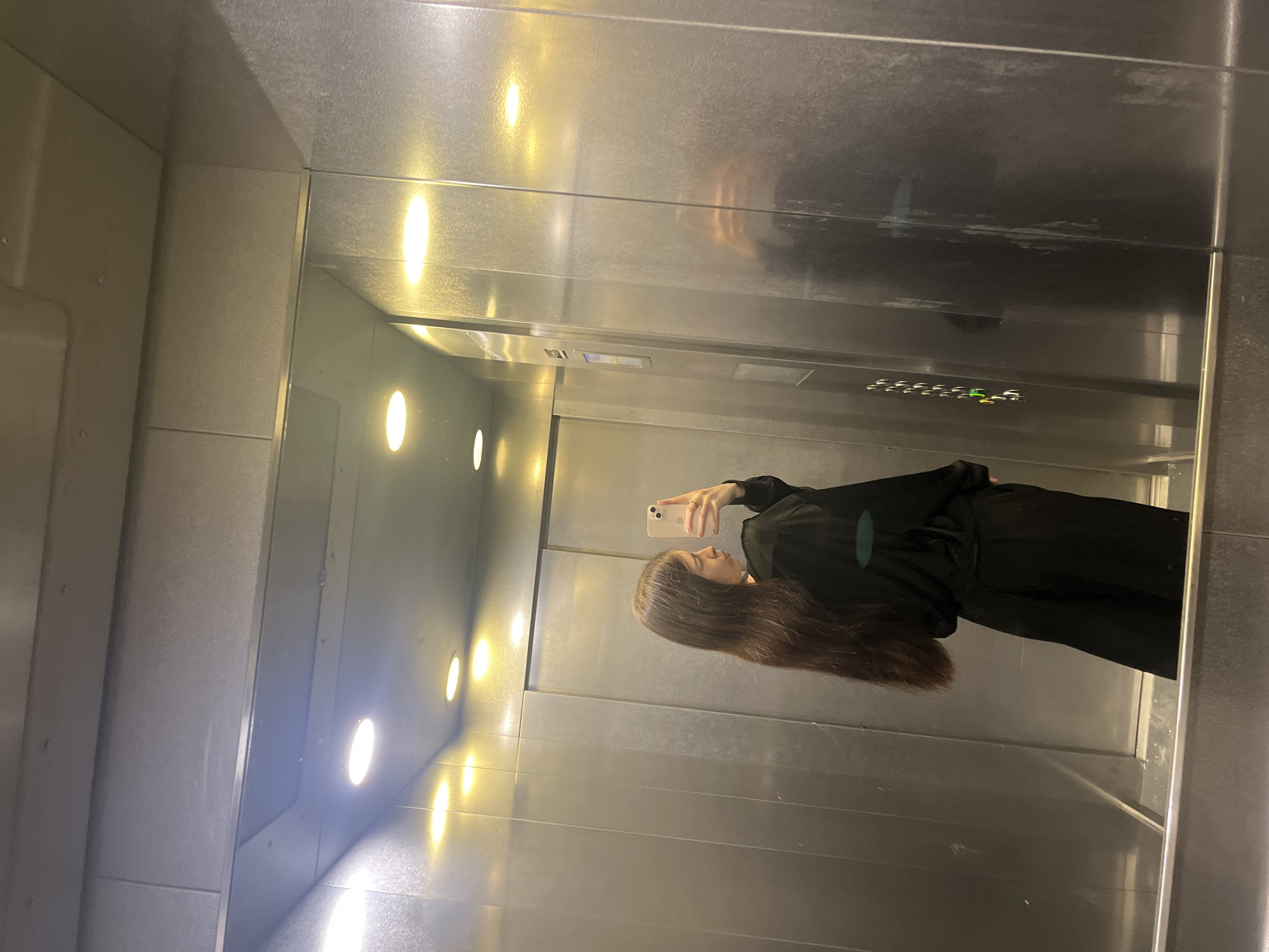 A person is taking a selfie in an elevator. The elevator has metal walls and a series of circular lights running vertically along the wall. The person is wearing a black outfit and has long brown hair. The reflection in the mirror shows the person holding a smartphone. It is not possible to determine the location in the city of Saint Petersburg, Russia, from the image.