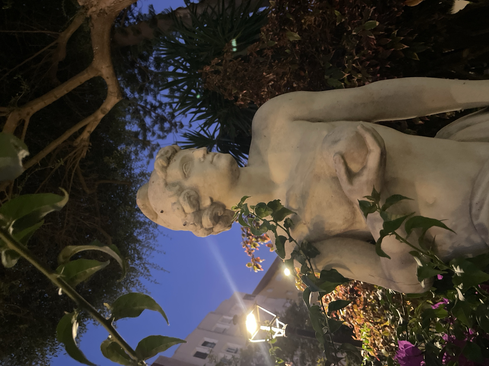 An outdoor statue of a person made of light-colored stone is situated in a garden area in La Línea de la Concepción, Spain, with a blue twilight sky visible in the background. The statue is positioned centrally in the frame and is partially obscured by foliage. The figure appears to be nude from the chest up, with its left hand resting on its chest, holding a round object. Green leaves and branches from an overhanging tree frame the top of the image, and smaller plants with purple flowers are visible at the lower left. A lamppost with a warm yellow light is visible to the left of the statue, and a building with windows is in the background, also illuminated. The overall scene suggests a garden or public space at dusk or early evening.