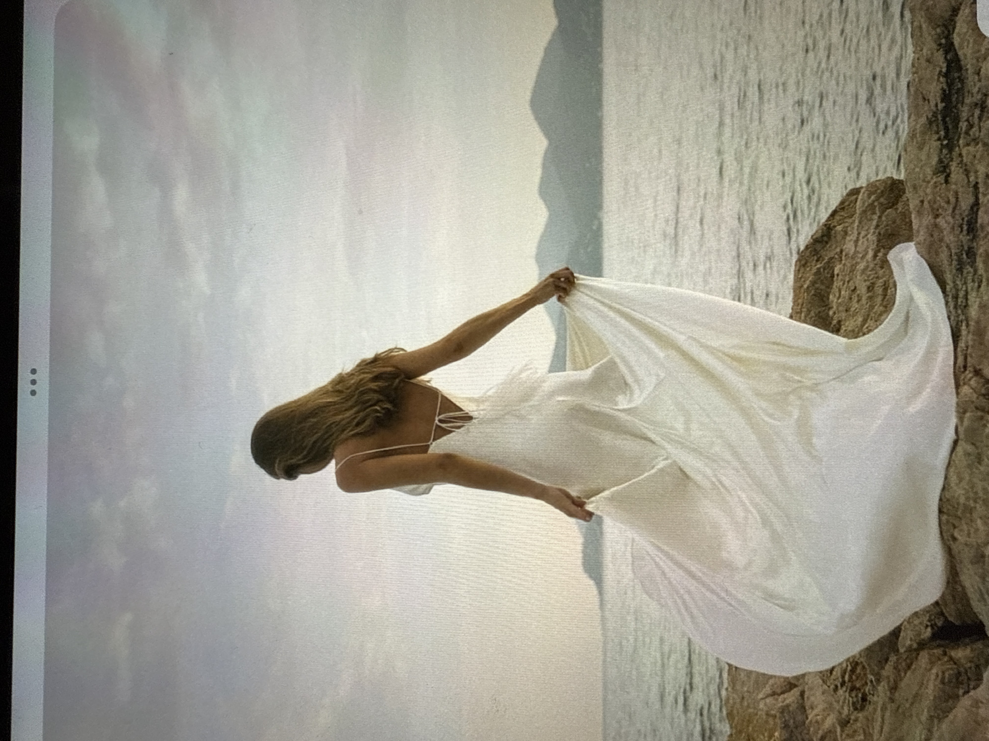 A woman stands on a rocky shoreline, facing away from the camera, her back to the viewer. She is wearing a flowing, white, spaghetti-strap dress that drapes to the ground and billows around her. Her hair is long and brown, falling down her back. The sea is visible in front of her, with a hazy, mountainous coastline in the distance. The sky above is overcast and pale. The location is not specified as Găvănoasa, Moldova.