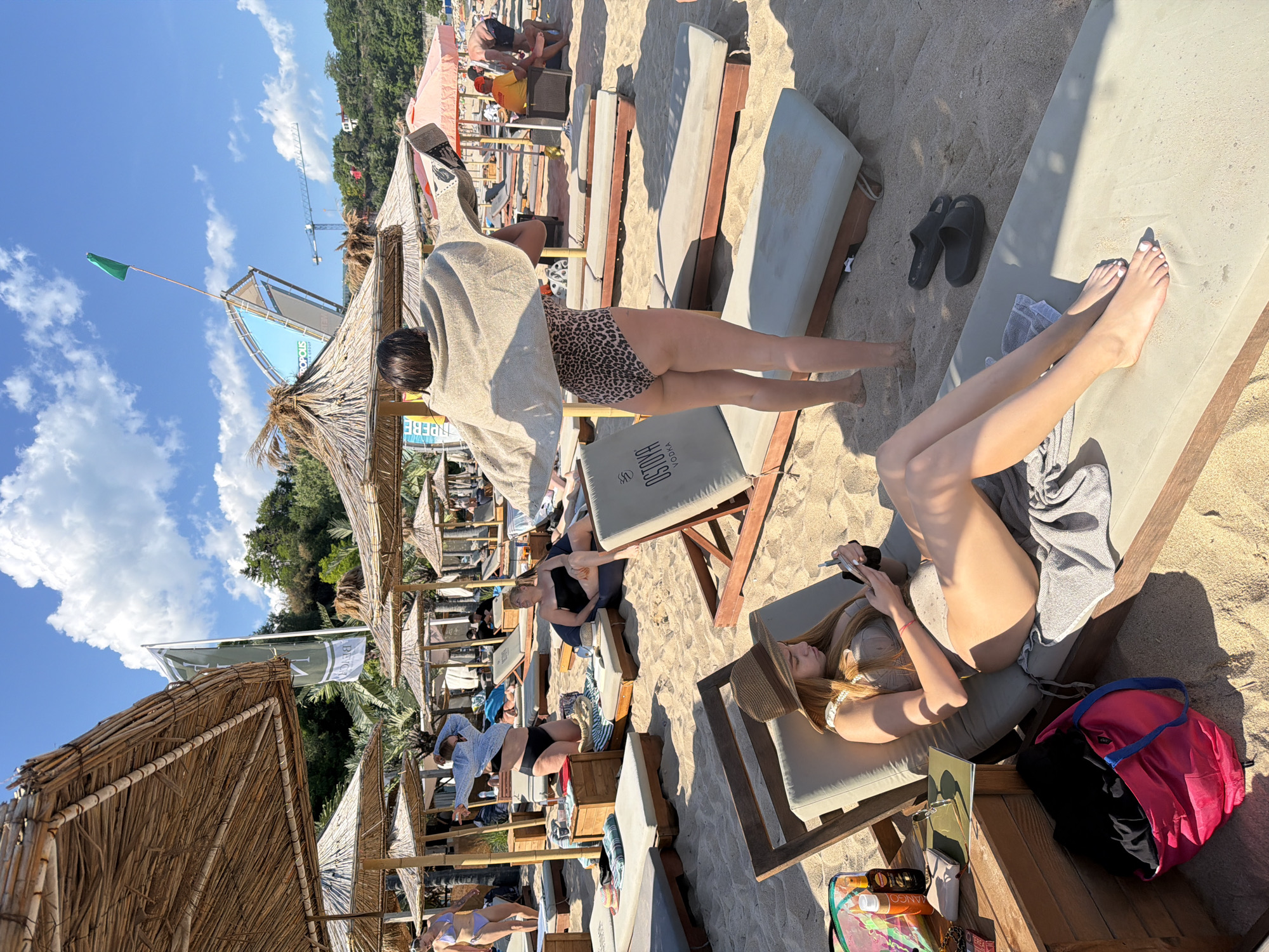 A crowded beach scene in Varna, Bulgaria, is depicted on a sunny day. The sand is covered with numerous beach chairs, most of which are occupied by sunbathers. Many straw umbrellas provide shade. In the foreground, a young woman wearing a sun hat and a light-colored bikini is reclining on a beach chair, looking at her phone. Next to her chair is a wooden side table with various items, including sunscreen, a wallet, sunglasses, and a book. A pink tote bag is also visible on the sand near her feet.
In the middle ground, a woman in a leopard-print swimsuit is standing and holding a beige towel over her shoulders. Behind her, other beachgoers are relaxing on lounge chairs. Further back, a person in a blue top and black bikini bottoms is adjusting their clothing. In the background, a large banner with the word "PROPOLIS" and graphics is visible. A green flag flies from a pole above a structure. Construction cranes are visible in the distance, suggesting ongoing development in the city. The sky is blue with scattered white clouds.