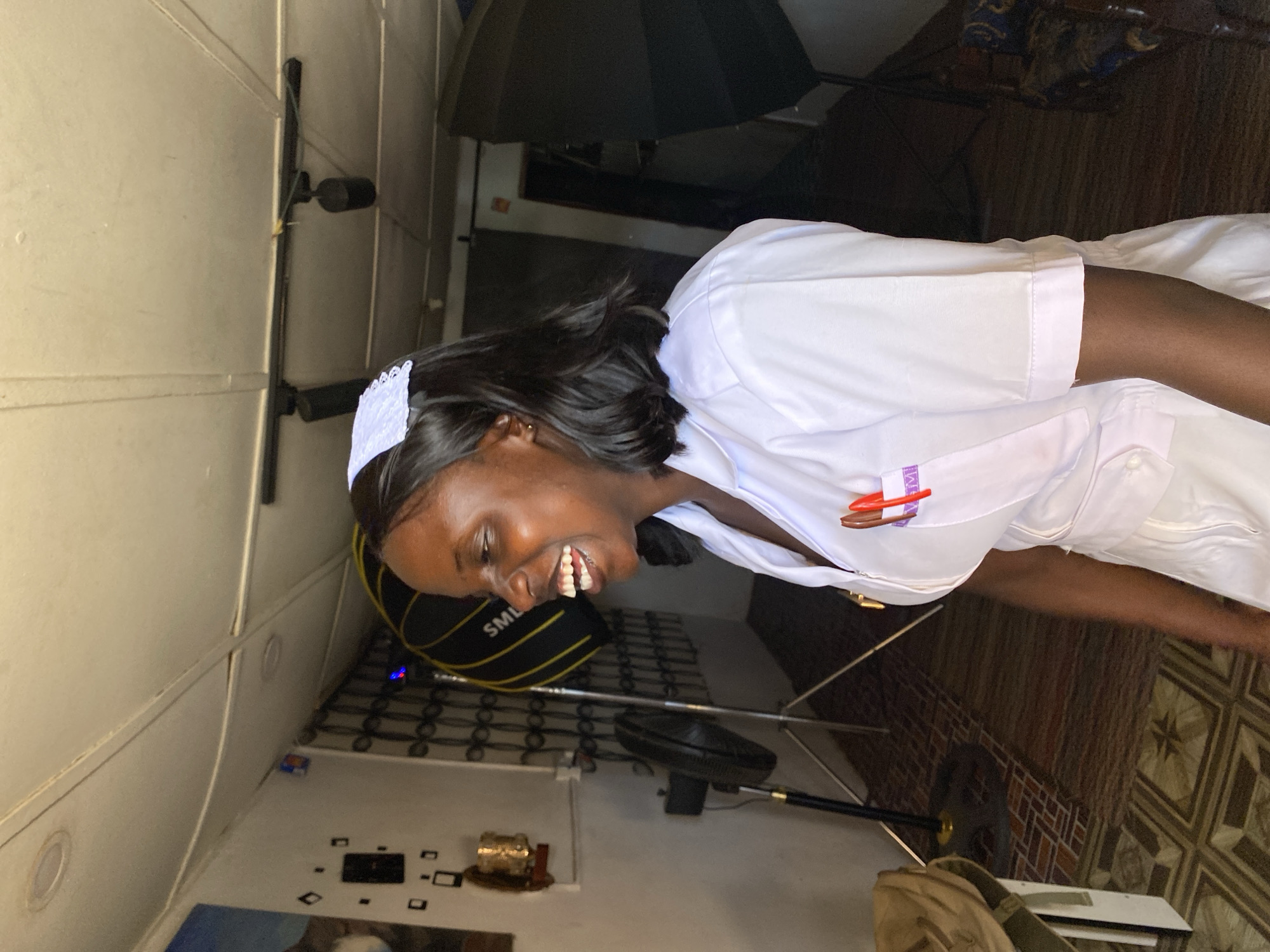 A woman wearing a white nurse uniform with a white cap is smiling and standing inside a room in Vom, Nigeria. A black umbrella and lighting equipment are visible behind her. A fan and a bag are also present in the room. The floor is tiled with a patterned design. A small shelf with objects on it is attached to the wall.