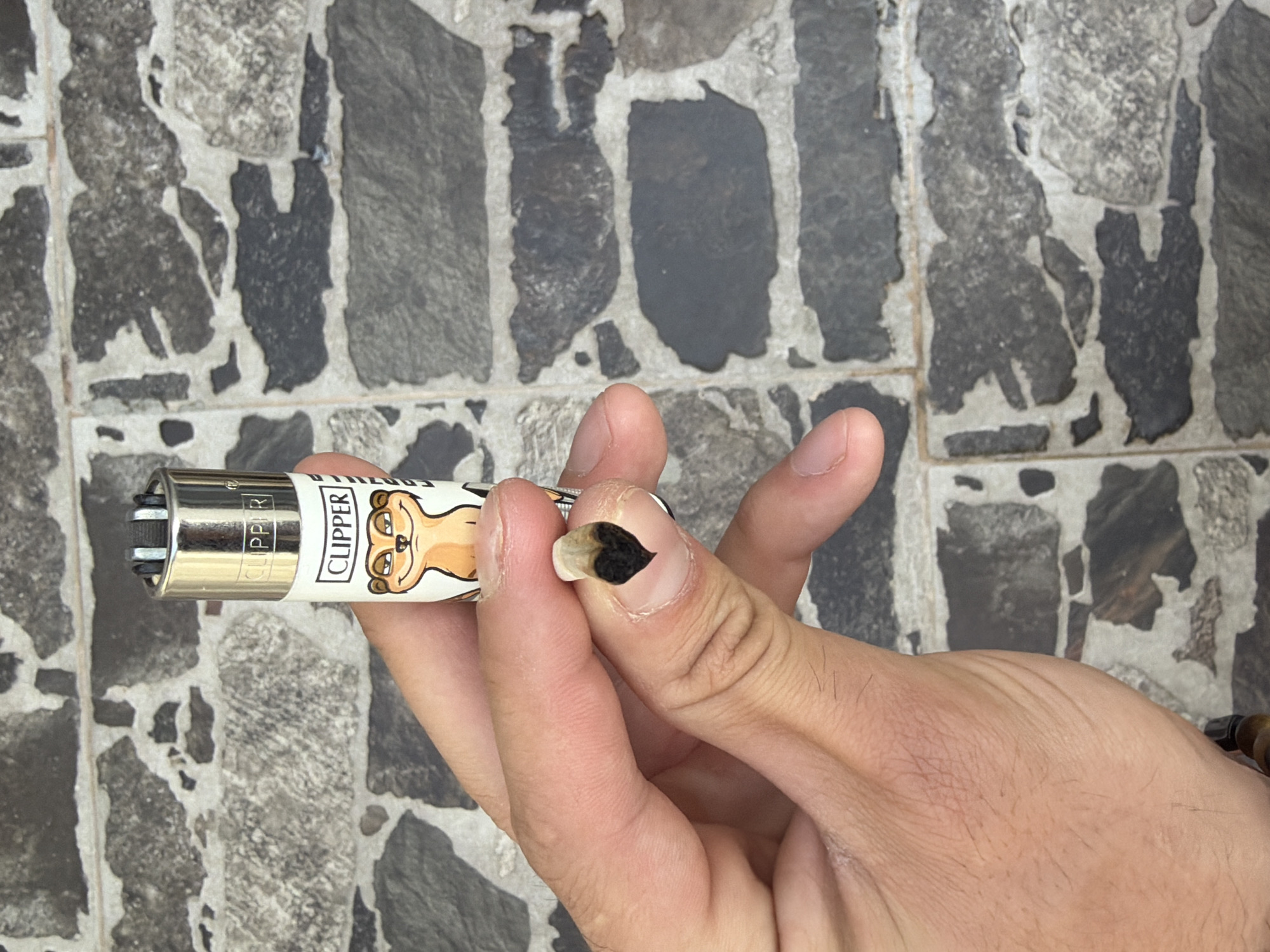 A close-up, top-down shot captures a person's hand holding a Clipper lighter and a burnt cigarette filter (roach) against a textured, mosaic-like tiled background, likely a floor or wall, in Plovdiv, Bulgaria.
The central subject is a light-skinned hand, held with the palm facing upwards. The thumb and index finger firmly grasp the burnt, dark-tipped cigarette filter, which appears heart-shaped due to its burnt edges. The other fingers are loosely curled. A beaded wooden bracelet is visible on the wrist in the lower right corner.
To the left of the hand, a cylindrical Clipper lighter is held, oriented horizontally. The lighter features a metallic silver top and a white body adorned with a cartoon illustration of a light brown, dog-like or meerkat-like animal with large eyes. The brand name "CLIPPER" is distinctly visible in black text on the white body of the lighter.
The background consists of irregular, dark grey and lighter grey stone-like tiles separated by light-colored grout, creating a varied, blocky pattern. The lighting appears even and natural, suggesting daylight without harsh shadows. No faces or other individuals are visible. The overall scene implies a moment of smoking, either just finished or about to begin, within a casual setting.