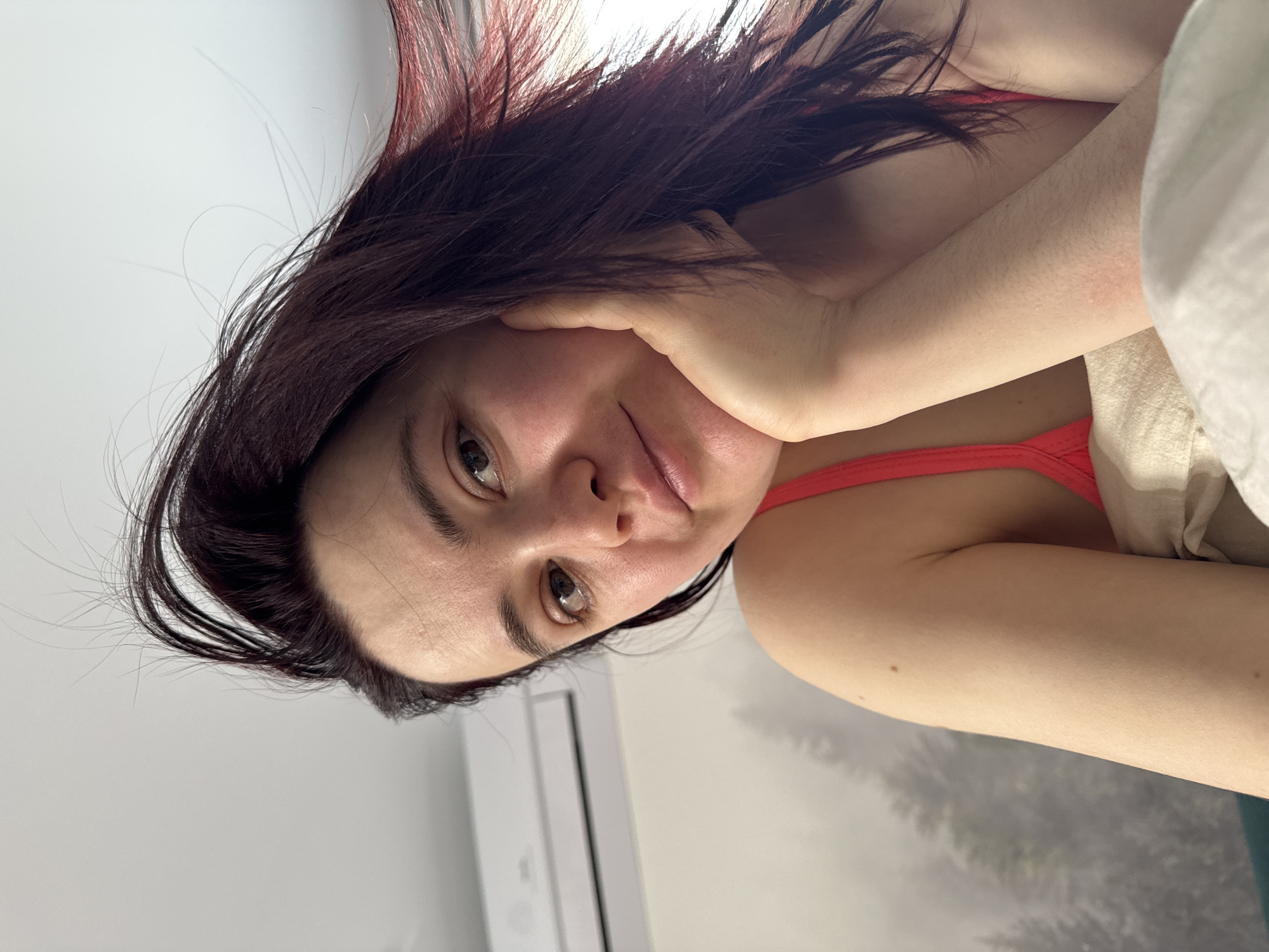 A woman with dark hair and fair skin is depicted in a selfie. She is lying down, with her head propped up by her hand. She is wearing a red tank top. Behind her, there is a white wall and a window air conditioning unit. The background appears to be a forest scene, possibly a wallpaper or a view from a window. The location is not identifiable as Izhevsk, Russia without additional context.