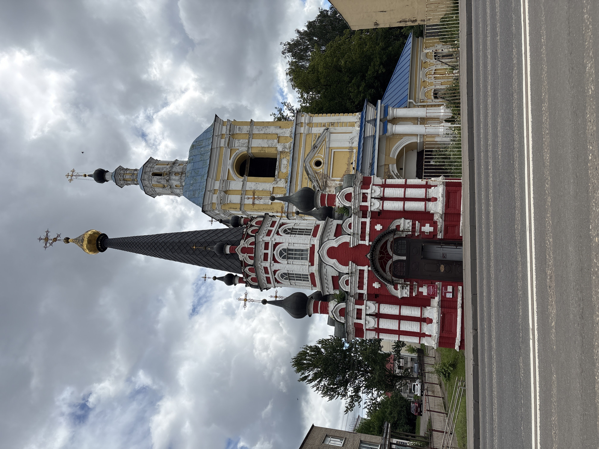 An exterior, eye-level, wide shot captures a red and white church with a tall, dark spire and a separate yellow bell tower with a blue roof, set against a cloudy sky. A paved road with white lane markings runs in the foreground, separating the viewer from a low, white metal fence enclosing the church grounds. Beyond the fence, grass and a few small white flowers are visible. To the left of the church, a brick building stands, and behind the church, trees and more buildings are partially visible. The church itself features multiple dark domes topped with crosses, and the red section has white decorative columns and arched windows. The bell tower is yellow with white trim and has arched openings. The overall scene is static and depicts a religious building within a town.