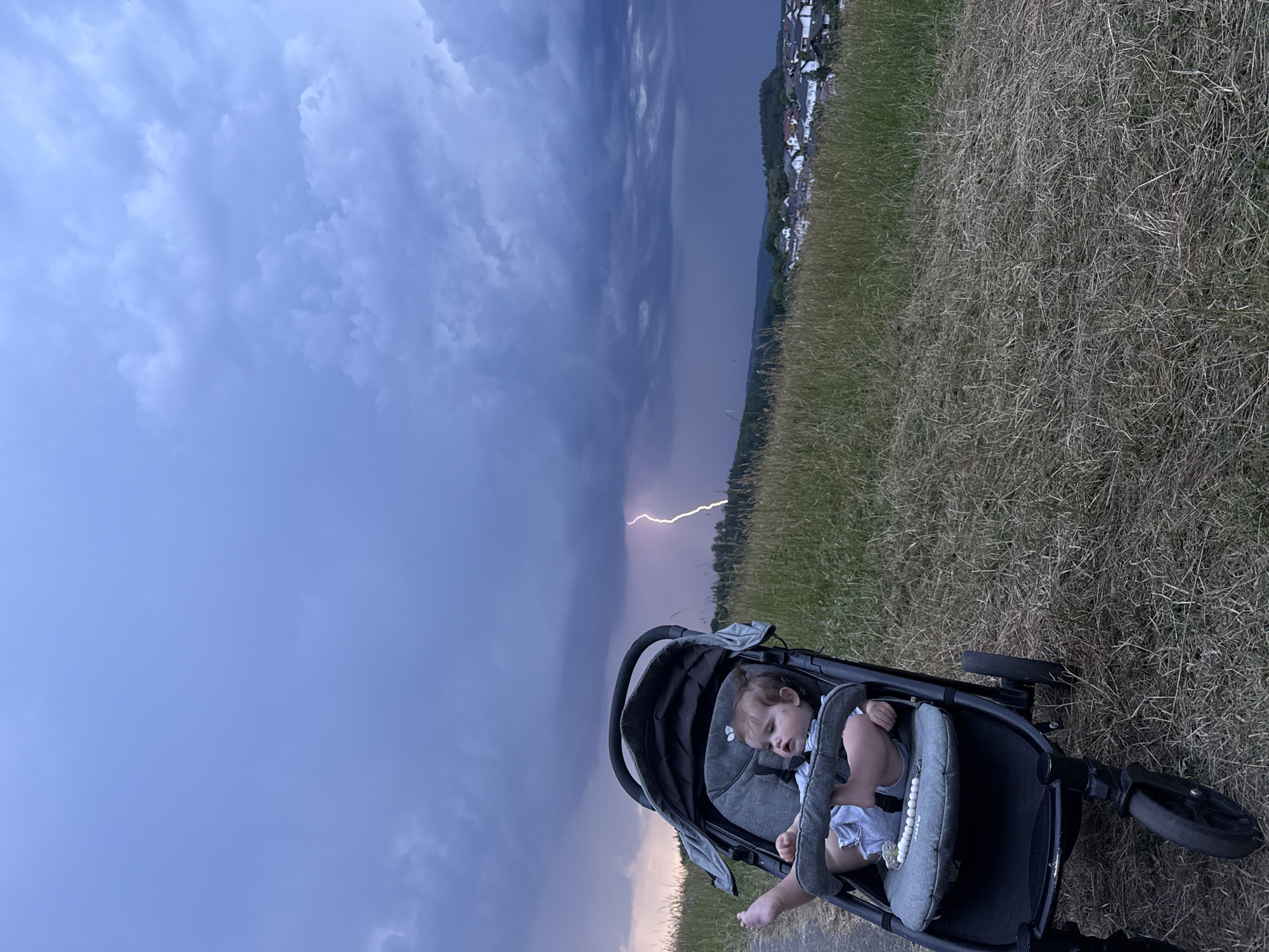 A baby is in a stroller on a grassy hill overlooking a town. The sky is dark and cloudy, with a lightning strike visible in the distance. The town, identified as Oberquembach, Germany, is nestled in the valley below, with houses and trees visible. The grass in the foreground is dry and brown, suggesting a hot or dry climate. The scene appears to be set during twilight or early evening, given the dim lighting and dark clouds.