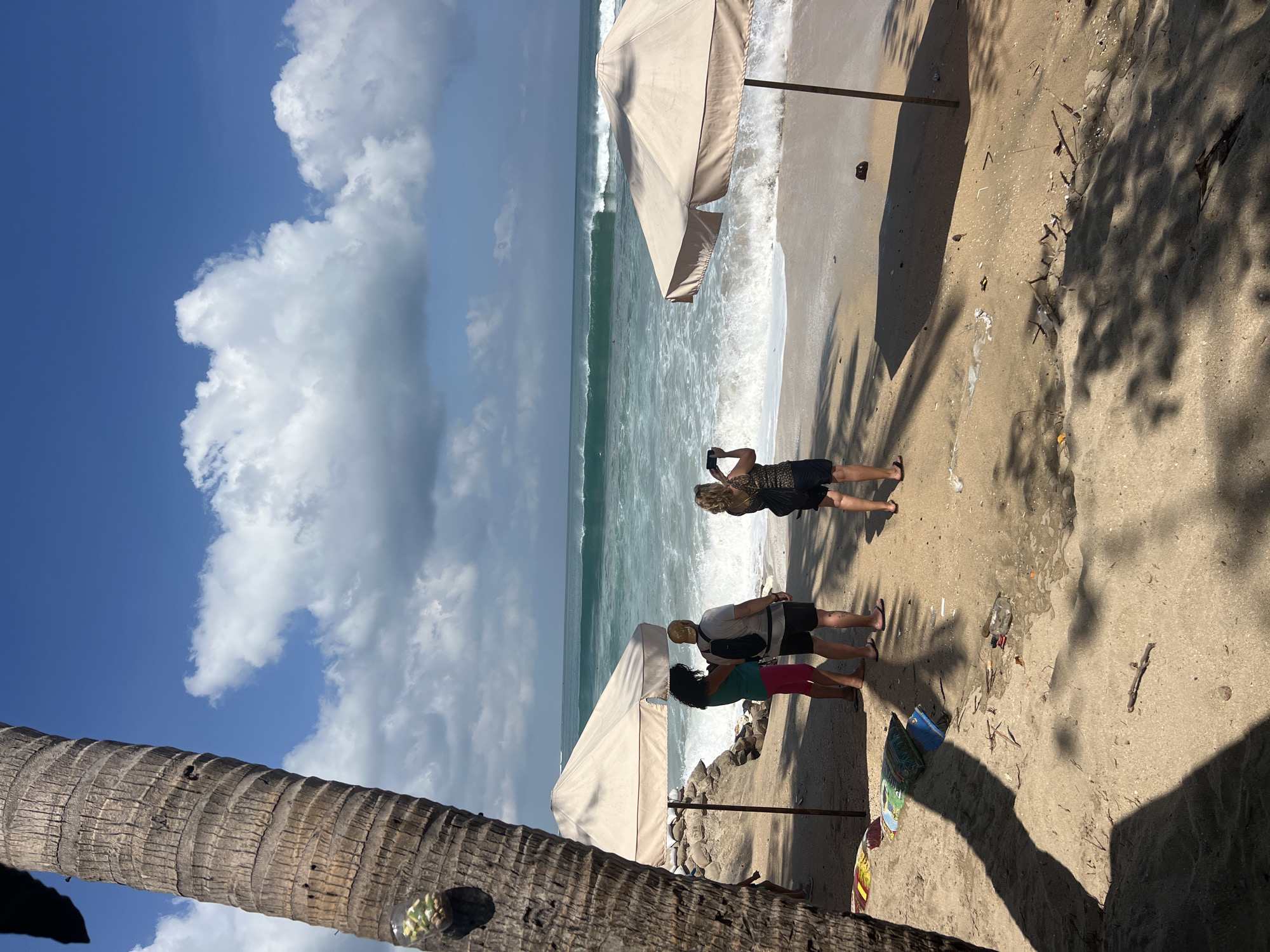 In Kuta, Indonesia, on a sunny day, a group of people are on a sandy beach near the ocean. The water is turquoise with white waves rolling in. Two beige umbrellas are set up on the sand. In the foreground, a large palm tree trunk is visible on the left side. Three people are standing near the water, facing the ocean. One person, wearing a green shirt and pink pants, is looking at the sea. Another person, in a grey shirt and black shorts, stands beside them, with a backpack. The third person, wearing a patterned shirt and black shorts, is taking a picture with their phone. Further back, two more individuals are partially visible. The sand is scattered with some debris, including a plastic bottle and dry leaves. Shadows of palm fronds are cast across the beach. The sky is blue with scattered white clouds.
