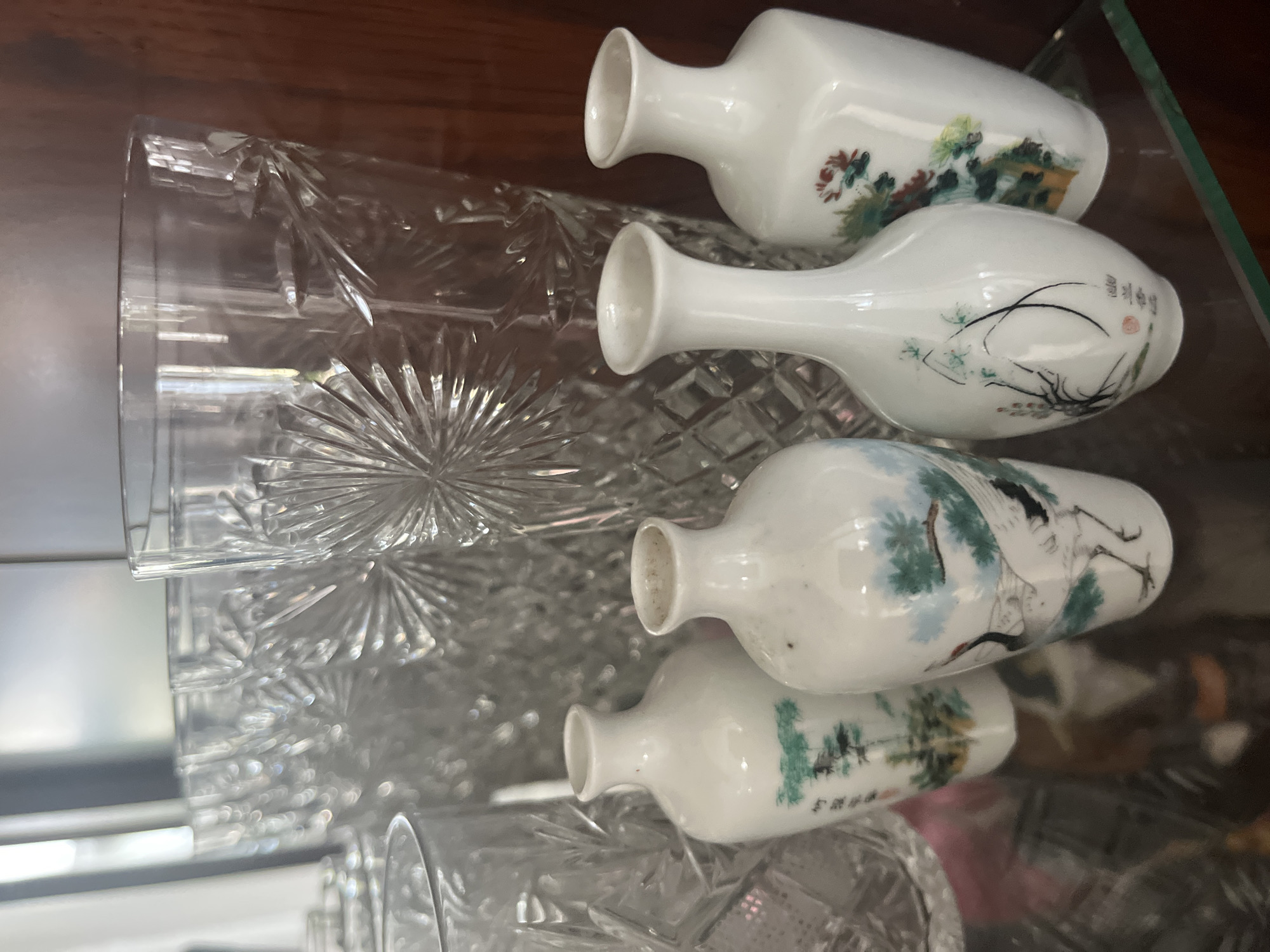 This is a close-up, eye-level, indoor shot of four small, white porcelain vases with painted designs, displayed on a glass shelf in what appears to be a display cabinet. The vases are arranged in a row, with two on the left and two on the right, slightly staggered in depth. The vase on the far left features a painted scene of trees. The second vase from the left depicts a crane amidst foliage. The third vase from the left has a delicate painting of branches and small red blossoms. The vase on the far right showcases a floral design with red and green elements.
Behind the vases, several tall, clear glass vessels with intricate cut-glass patterns are visible. These glasses are partially obscuring the background, which appears to be the interior of a wooden cabinet. The lighting is soft, creating reflections on the glass surfaces. The overall impression is one of a curated collection of decorative items.
The image is not identifiable as being located in Vladimirovka, Russia.