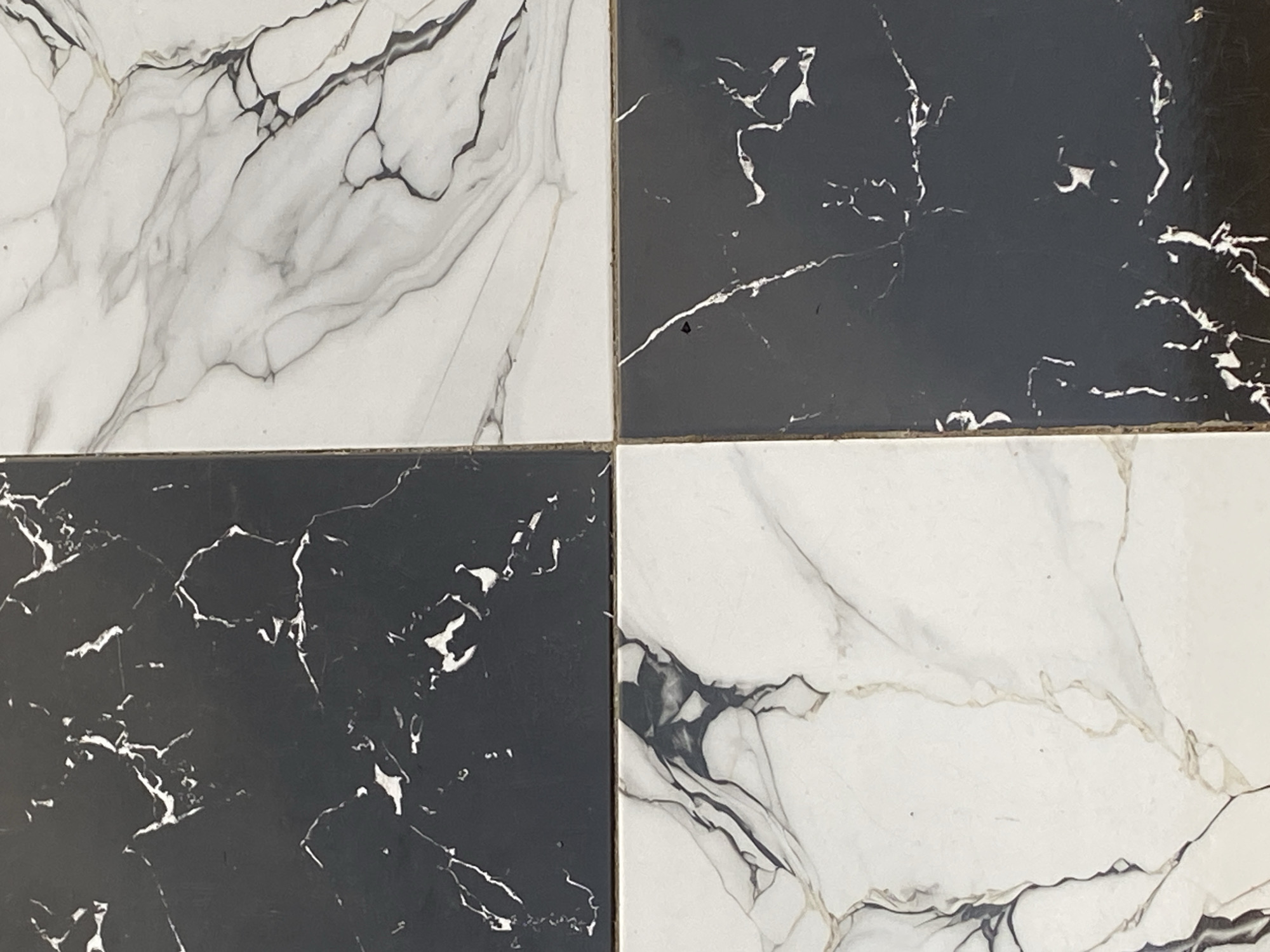 The image shows a close-up, overhead view of four square tiles arranged in a 2x2 grid. Two tiles are black with white veining, and two are white with black and gray veining, mimicking the appearance of marble. The tiles are positioned on a flat, presumably floor or countertop surface. No people, actions, or interactions are visible. There are no discernible environmental features that would allow for location identification within Jauro Yinu, Nigeria, or any other city. The scene is purely decorative.