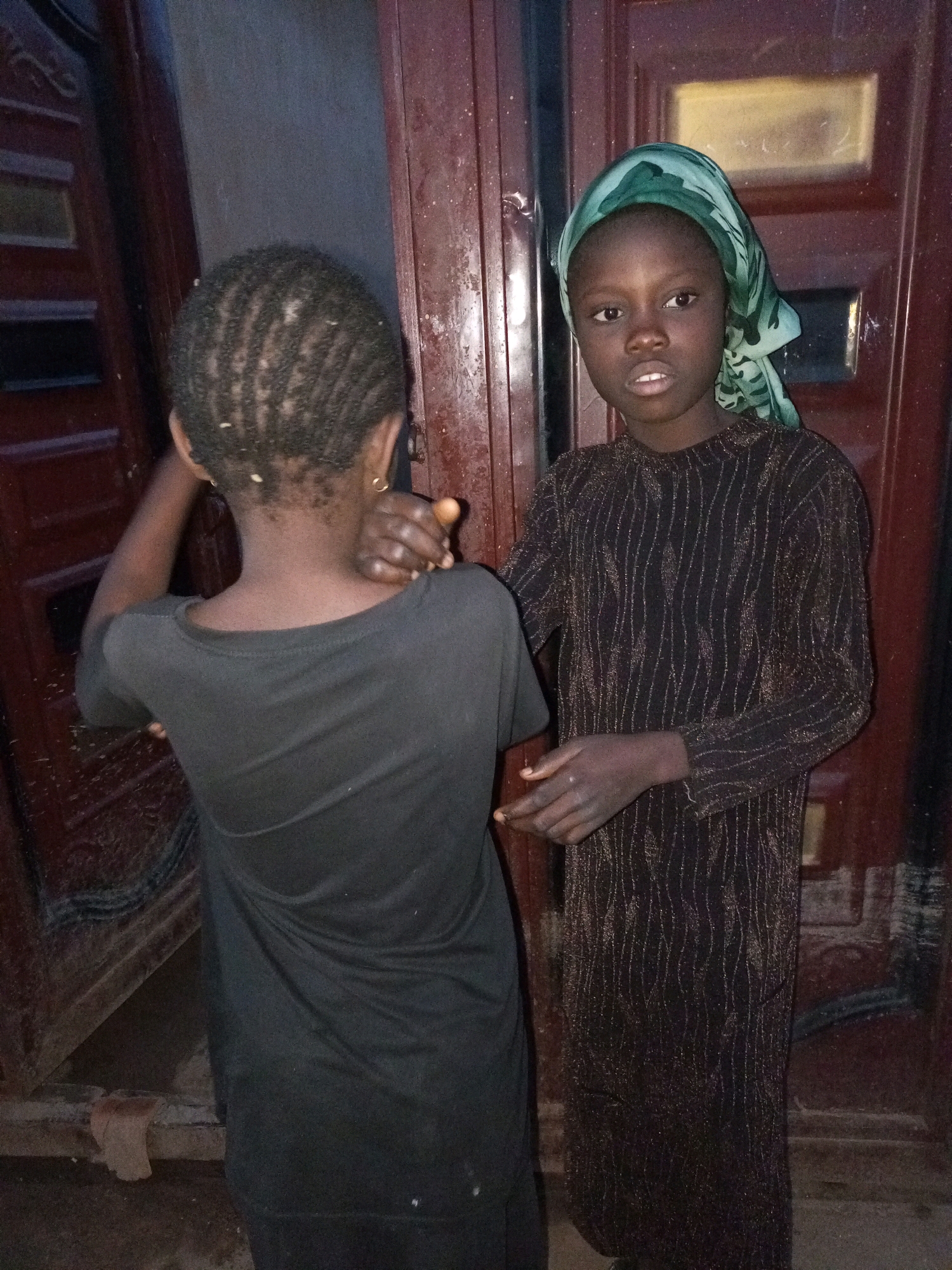 In Tempure, Nigeria, two young individuals stand in front of a dark wooden doorway. One person, facing away from the camera, wears a simple black t-shirt. Their hair is styled in small braids, and they have a gold hoop earring in their visible ear. Their left arm is bent, with their hand resting on their shoulder. The second individual, facing the camera, is adorned with a green headscarf and a long-sleeved black dress with shimmering vertical stripes. Their expression is neutral as they look towards the viewer. The background consists of a weathered wooden door with vertical panels and a rectangular inset, possibly a window, filled with a yellowish light. The ground beneath them appears to be dirt.