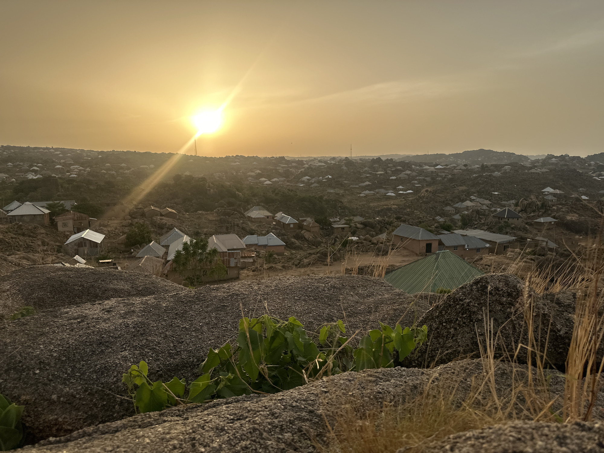 The image shows a landscape with a view of a residential area situated among rocky terrain, possibly on the outskirts of Jos, Nigeria. The scene is bathed in the warm light of the setting sun, which is positioned prominently in the upper center of the frame.
Numerous buildings are scattered across the landscape, nestled between the rocks. The structures appear to be single-story dwellings, with a variety of roofing materials visible, ranging from corrugated metal to what appears to be traditional thatch.
In the foreground, large, coarse rocks dominate the view. Patches of vegetation, including small green plants and dry grass, grow amidst the rocks.